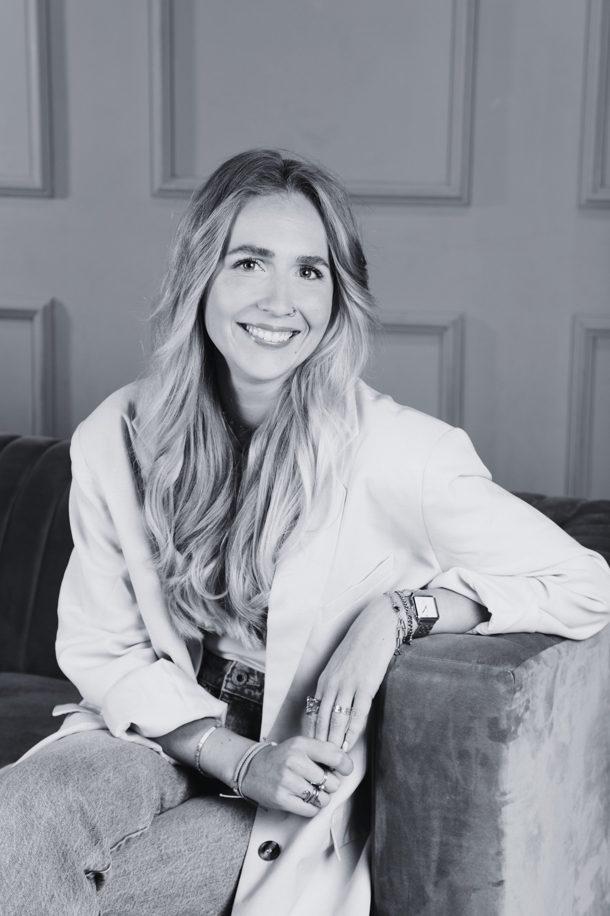 A smiling woman with long wavy hair sitting on a couch in a room with paneled walls, wearing a light-colored blazer, jeans, and jewelry.