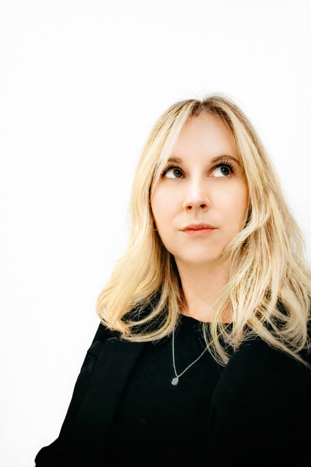 A blonde woman looking upwards with a thoughtful expression, wearing a black top and a necklace, against a white background.