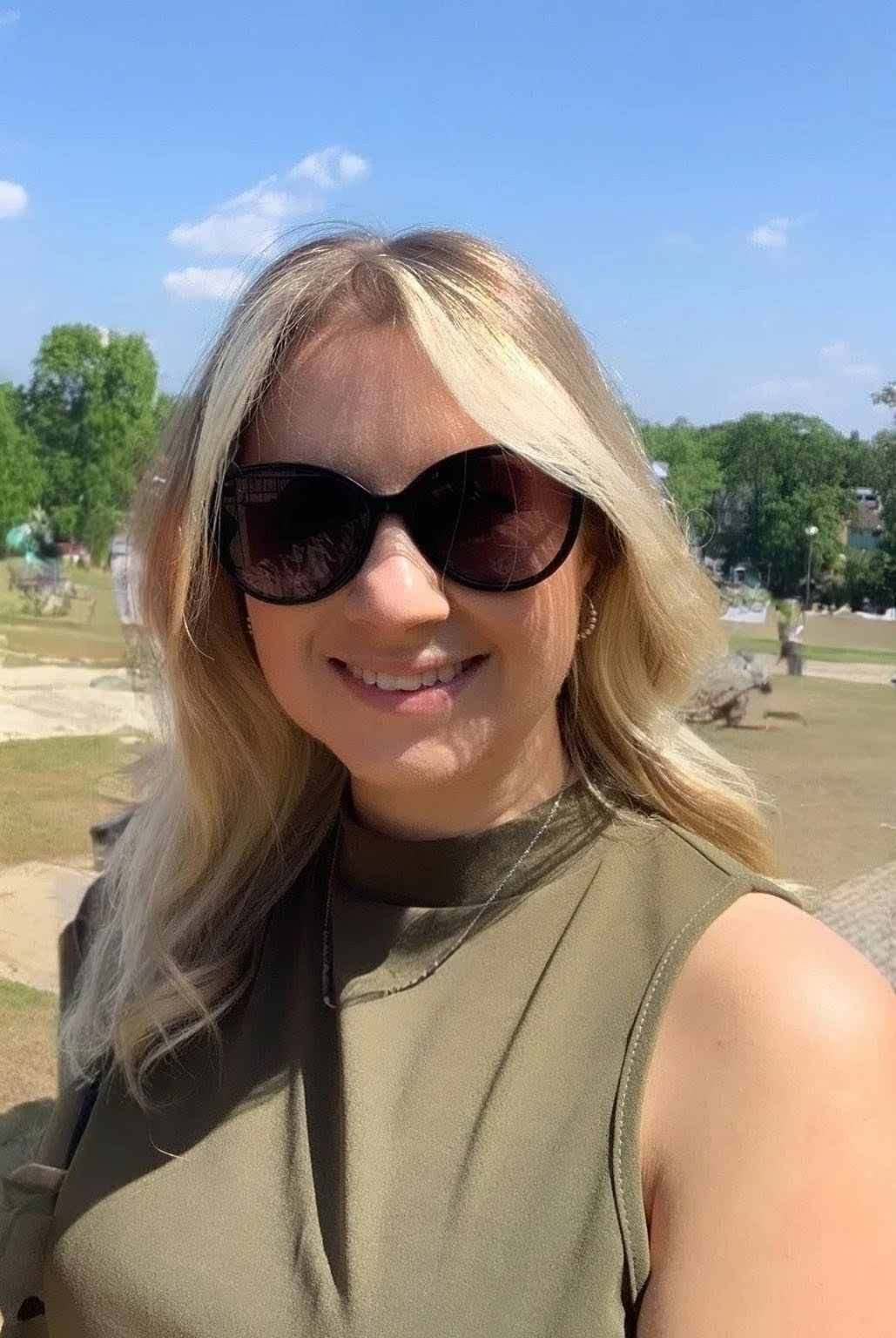 A woman with blonde hair wearing black sunglasses and a sleeveless olive green top, smiling outdoors on a sunny day with green trees and a park in the background.