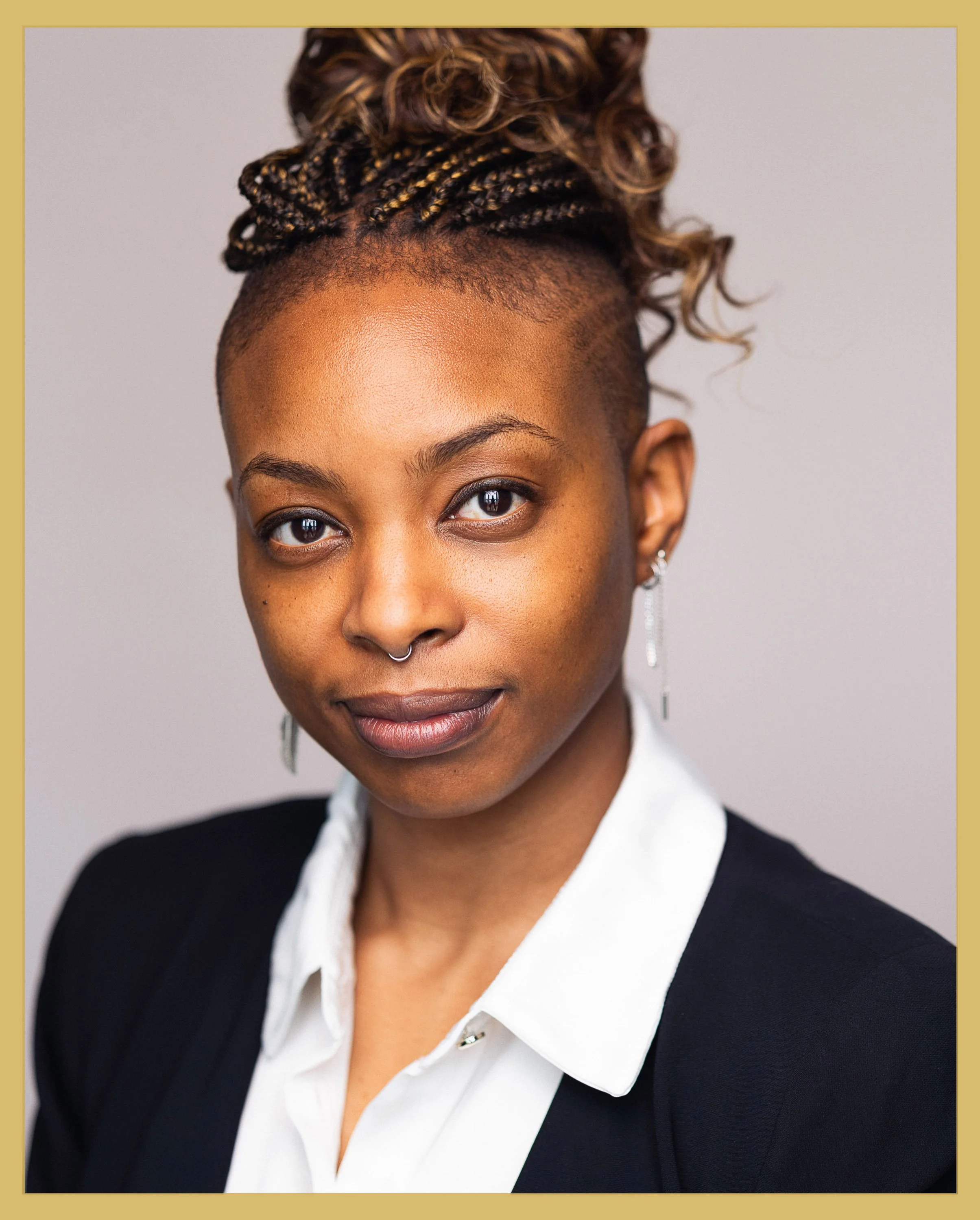 Portrait of a woman with styled hair, wearing a white collared shirt and black blazer, with earrings and a septum piercing, smiling subtly against a plain background.
