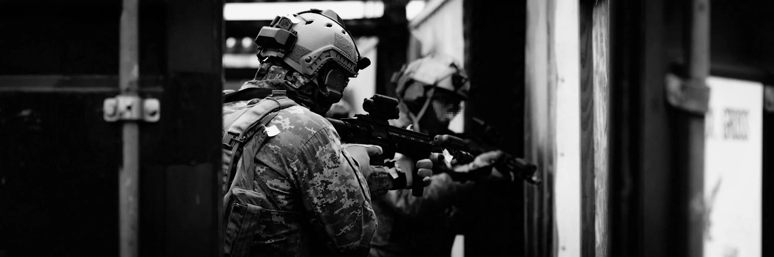 Two soldiers in camouflage uniforms and helmets aiming rifles through a narrow opening in a wooden structure, in black and white.