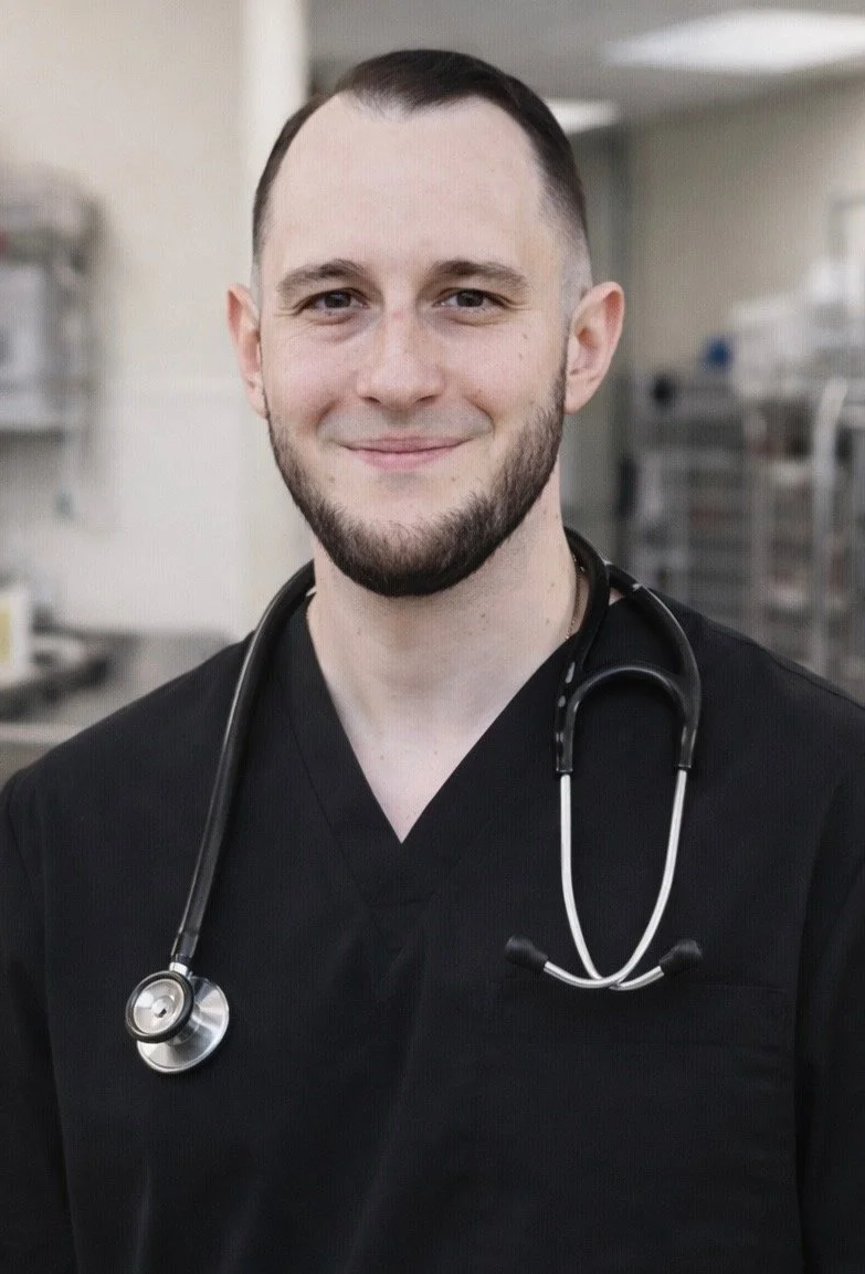 A male healthcare professional smiling, wearing black scrubs and a stethoscope around his neck, in a medical setting with shelves and medical equipment in the background.