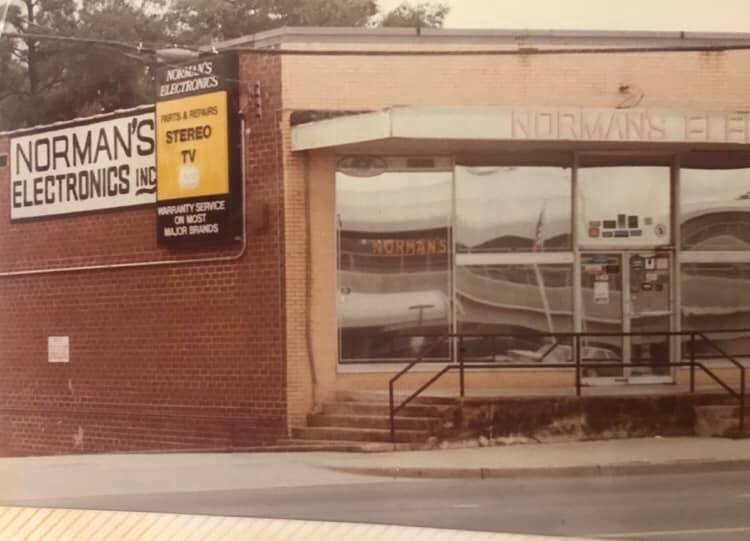 Exterior view of Norman's Electronics store with a large sign, entrance with glass doors and windows, and a ramp leading up to the door.