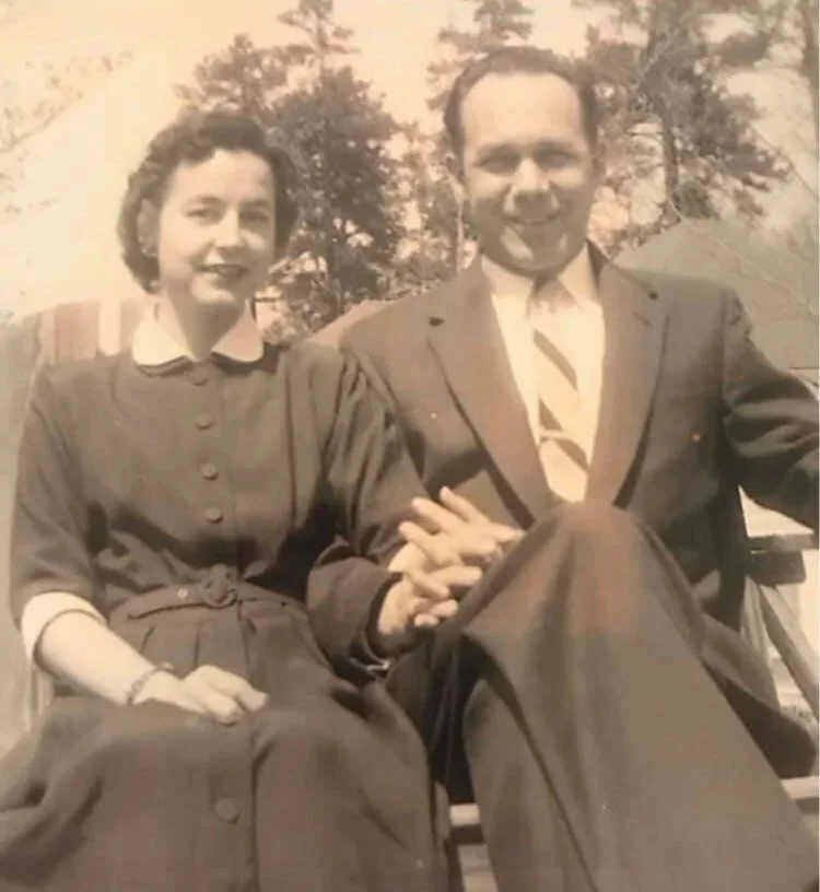 A black and white photo of a man and woman sitting together outdoors, holding hands. The woman has short wavy hair and is wearing a buttoned dress with a white collar, while the man has short hair and is dressed in a suit with a tie. They are smiling and appear happy, with trees in the background.