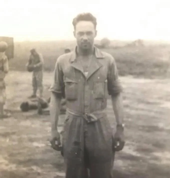 Black and white photo of a man in military uniform holding kettlebells in both hands, standing outdoors with other soldiers in the background.
