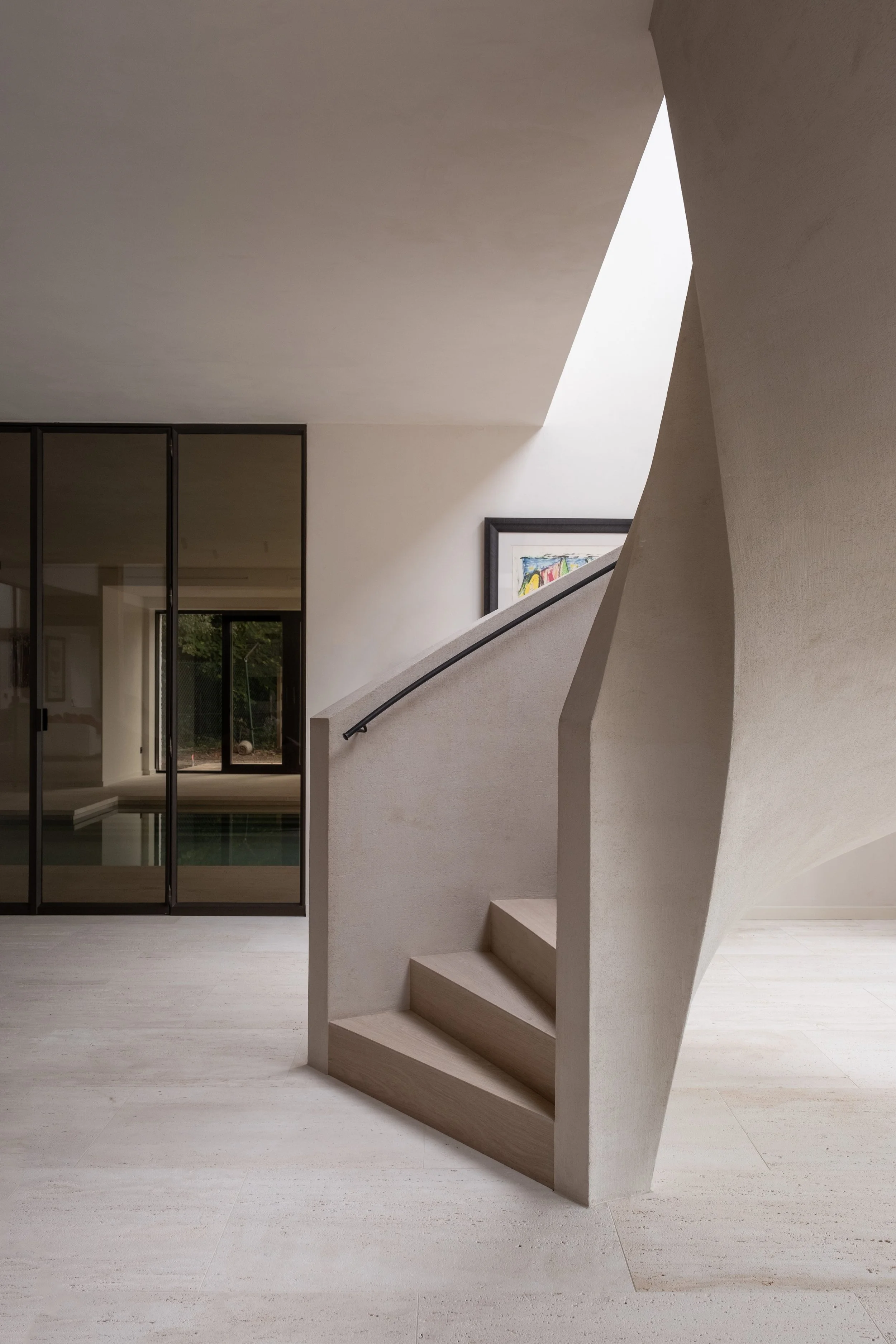 Minimalistisch interieur met trappenhuis, beige muren en vloeren, een kleurrijk kunstwerk, en een glazen schuifdeur die uitkomt op een tuin met een zwembad.