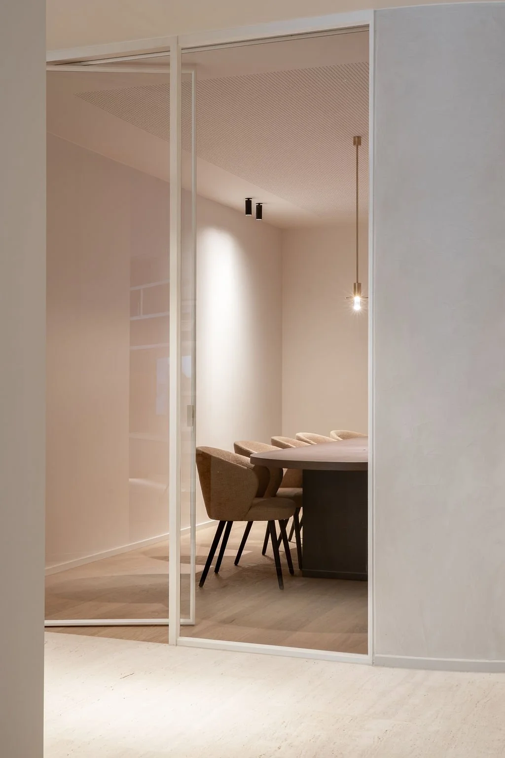 Modern eetkamer met ronde tafel en vier beige eetstoelen, scheidingswand met glazen deur, zachte verlichting, houten vloer en lichte muren.