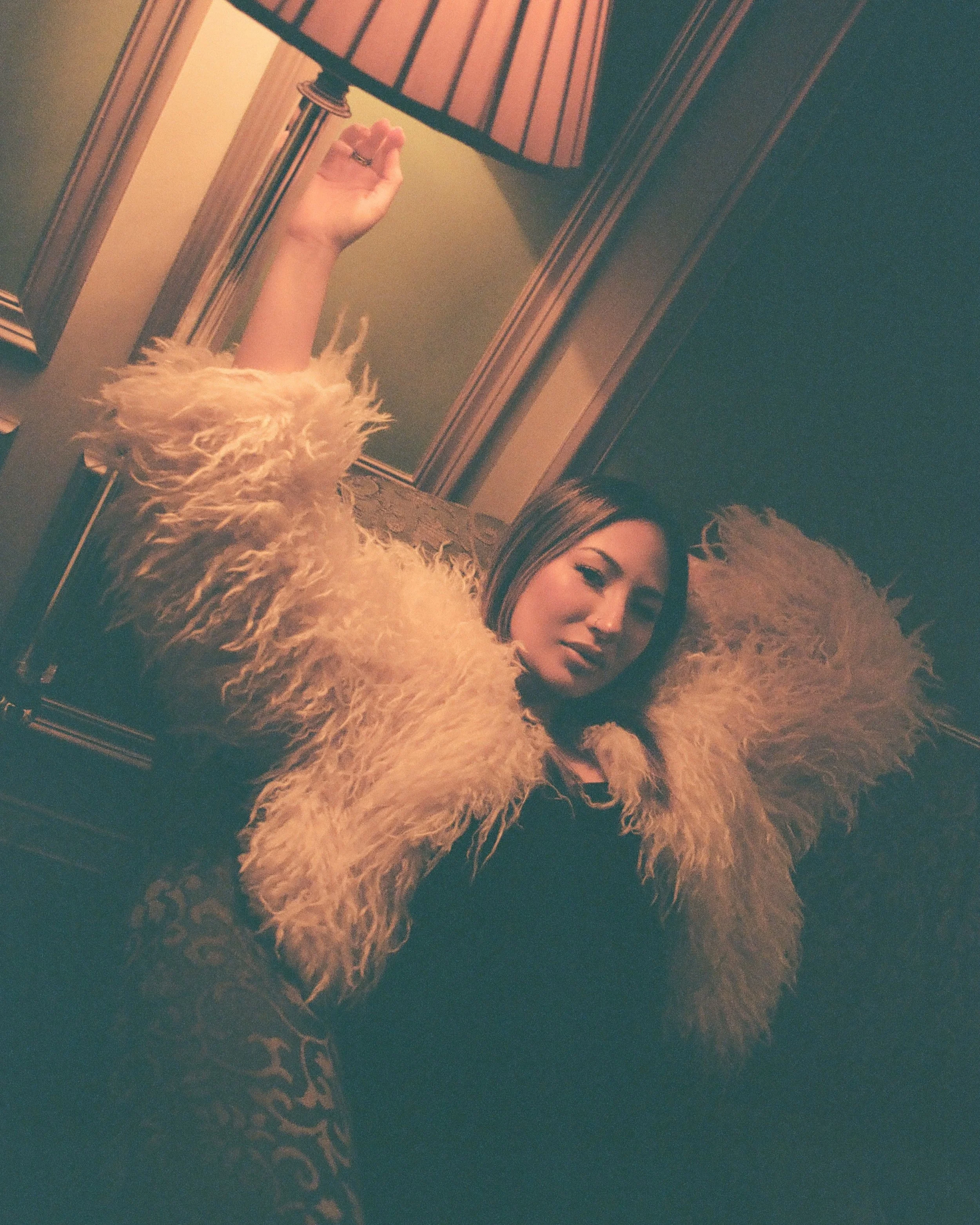 A woman leaning back on a couch wearing a beige furry coat, with a lamp on a wooden side table and decorative wallpaper visible in the background.