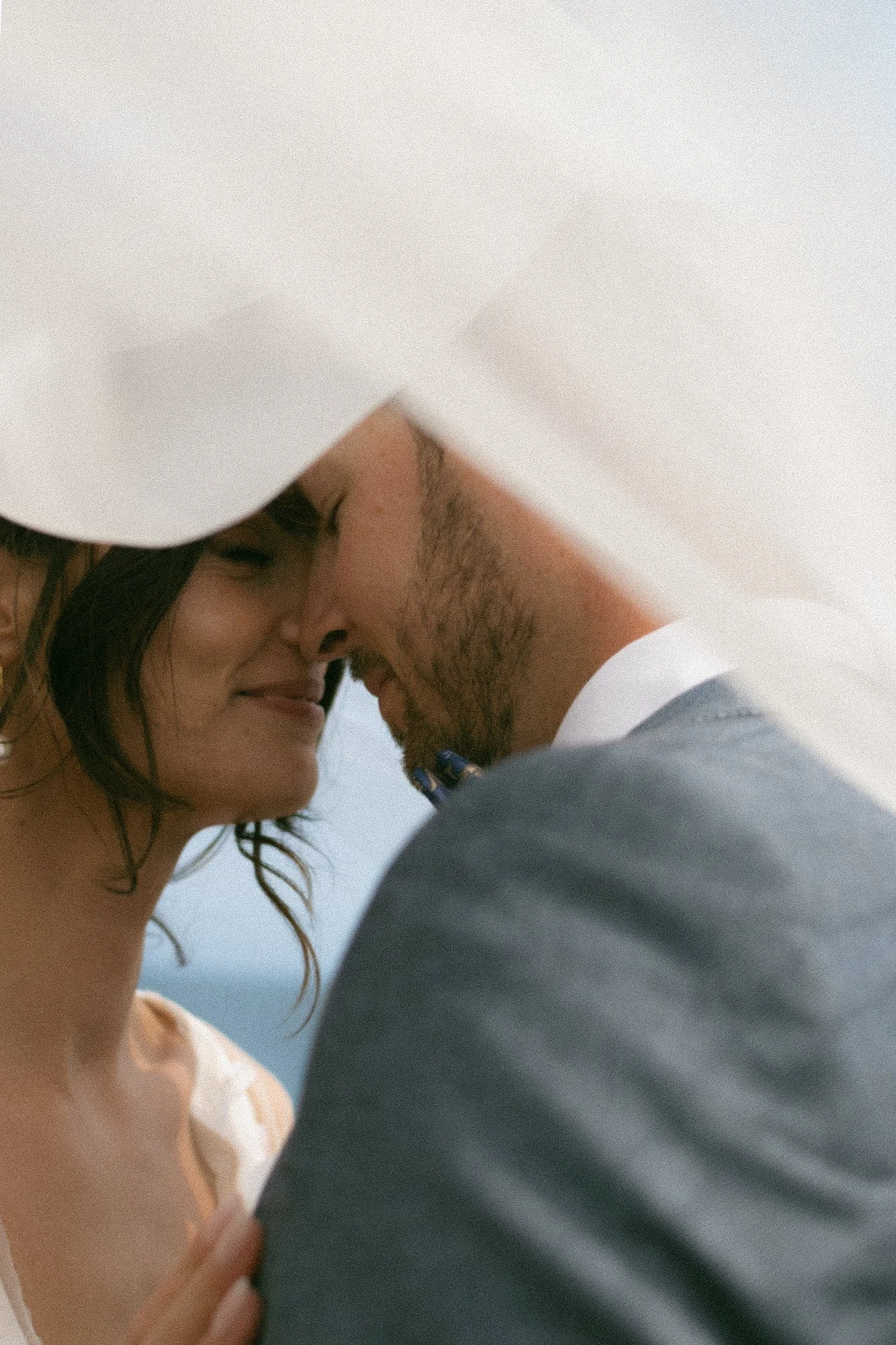 A close-up of a couple's faces softening as they touch foreheads and smile, with the woman having dark, wavy hair and the man with a beard and short hair. They are outdoors, possibly at a beach or open sky background.