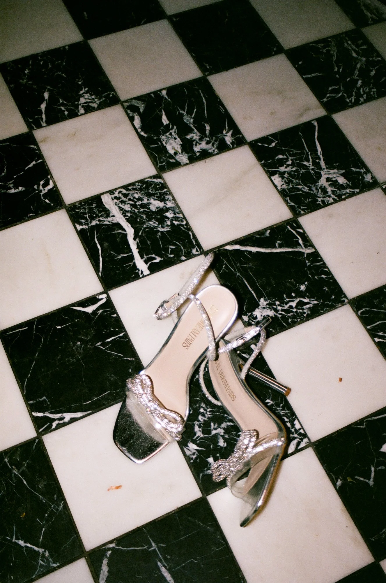 A pair of silver high-heeled sandals with rhinestone embellishments on a black and white checkered marble floor.