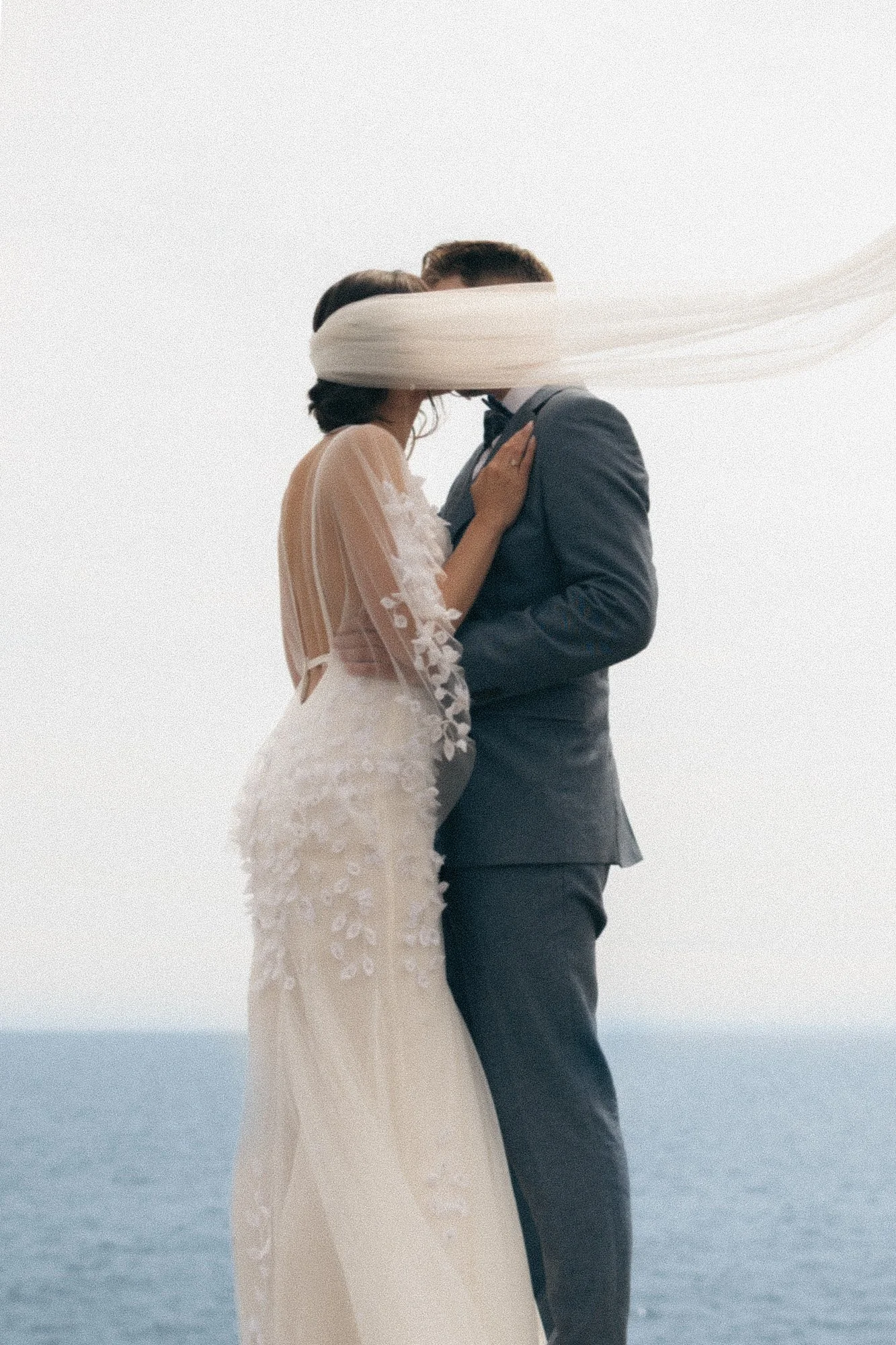 A newlywed couple standing on a beach, embracing, with the bride wearing a lace wedding dress and the groom in a suit, their faces covered with a stream of flowing white fabric.