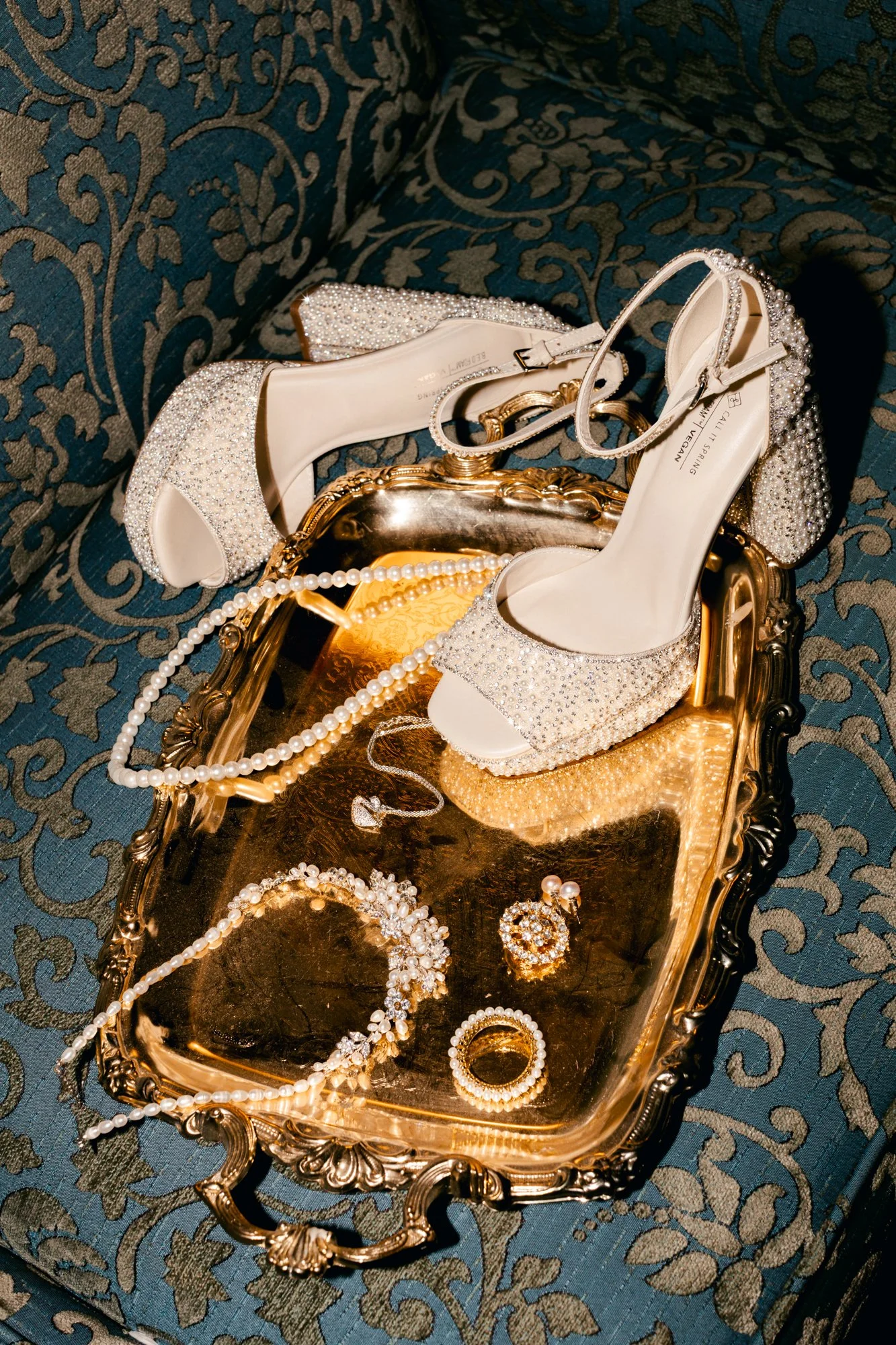 Jewelry and a pair of white high-heeled shoes with pearl embellishments placed on a gold tray, set on a decorative blue and gold patterned fabric.
