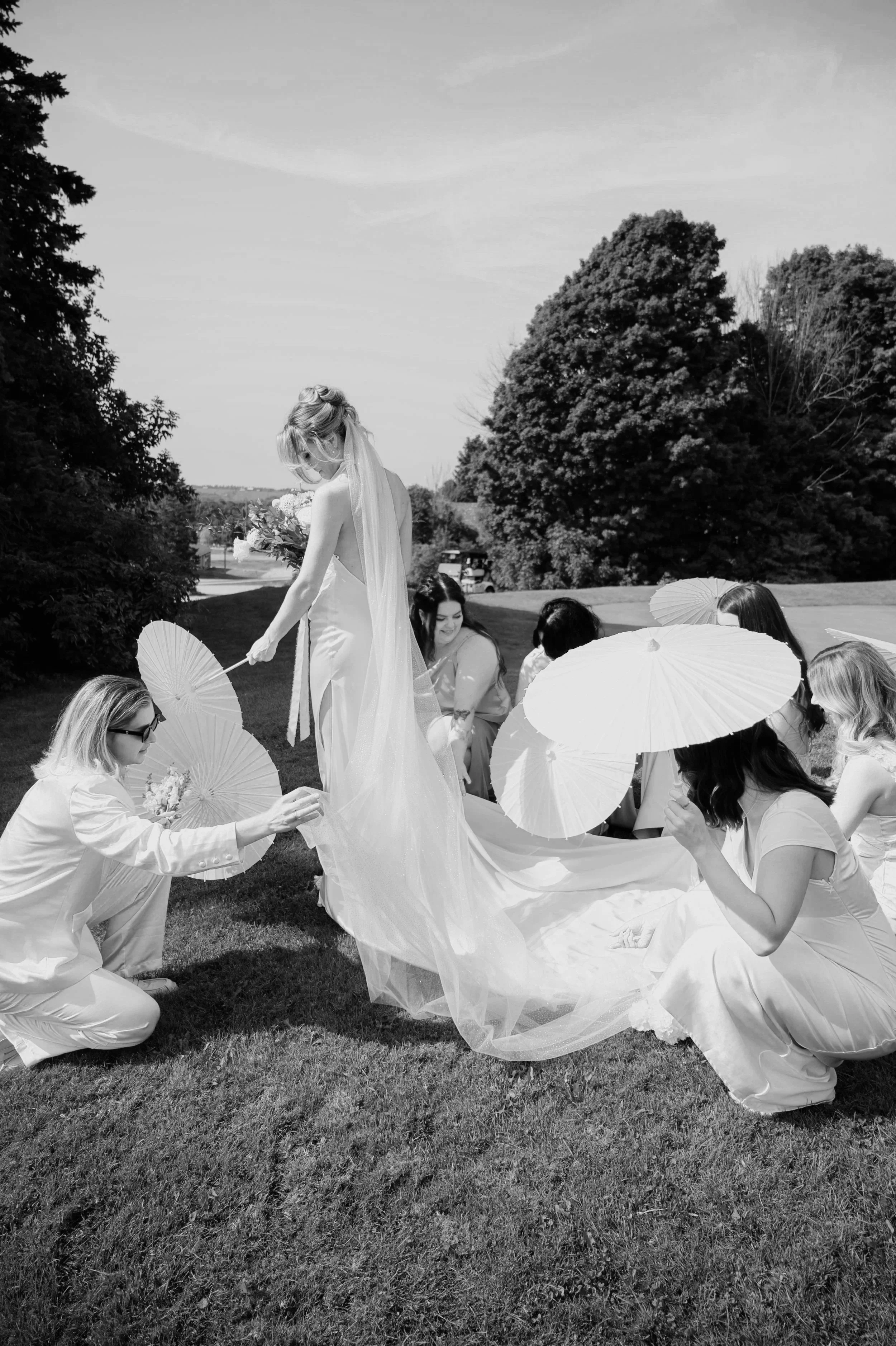 A bride in a wedding dress standing outdoors on the grass, surrounded by women in dresses holding parasols, with trees and sky in the background.