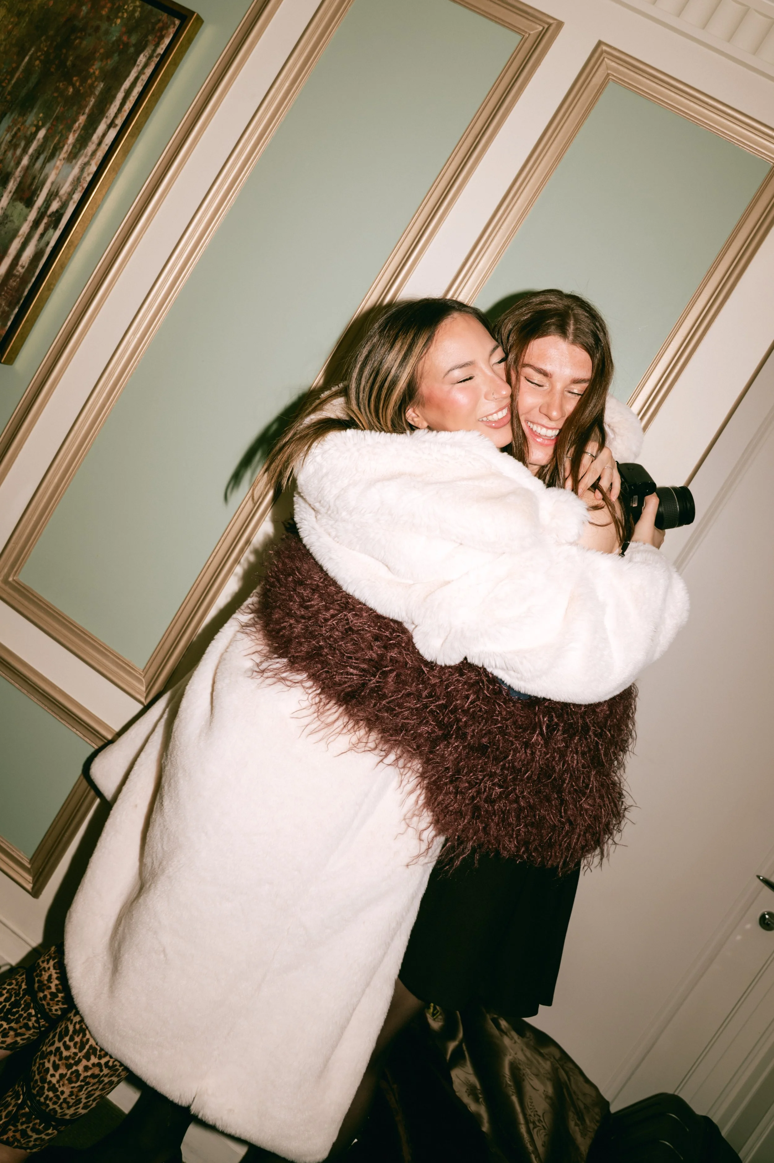 Two women hugging and smiling, standing in front of a green wall with gold-trimmed mirror frames, one holding a camera, both wearing warm clothing.