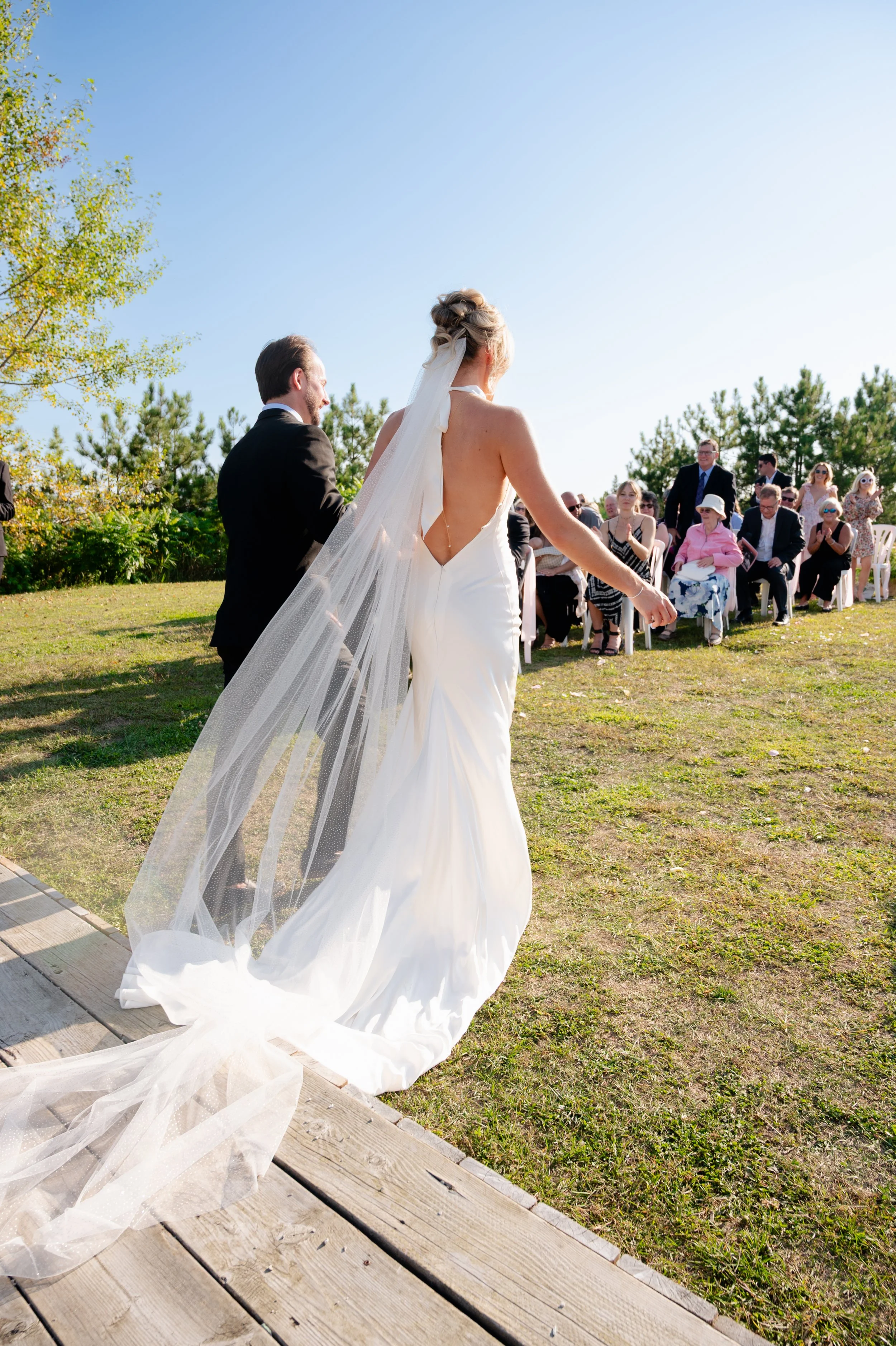 Bride and groom walking down a wooden aisle outdoors, guests seated on chairs watching, trees and clear sky in the background.