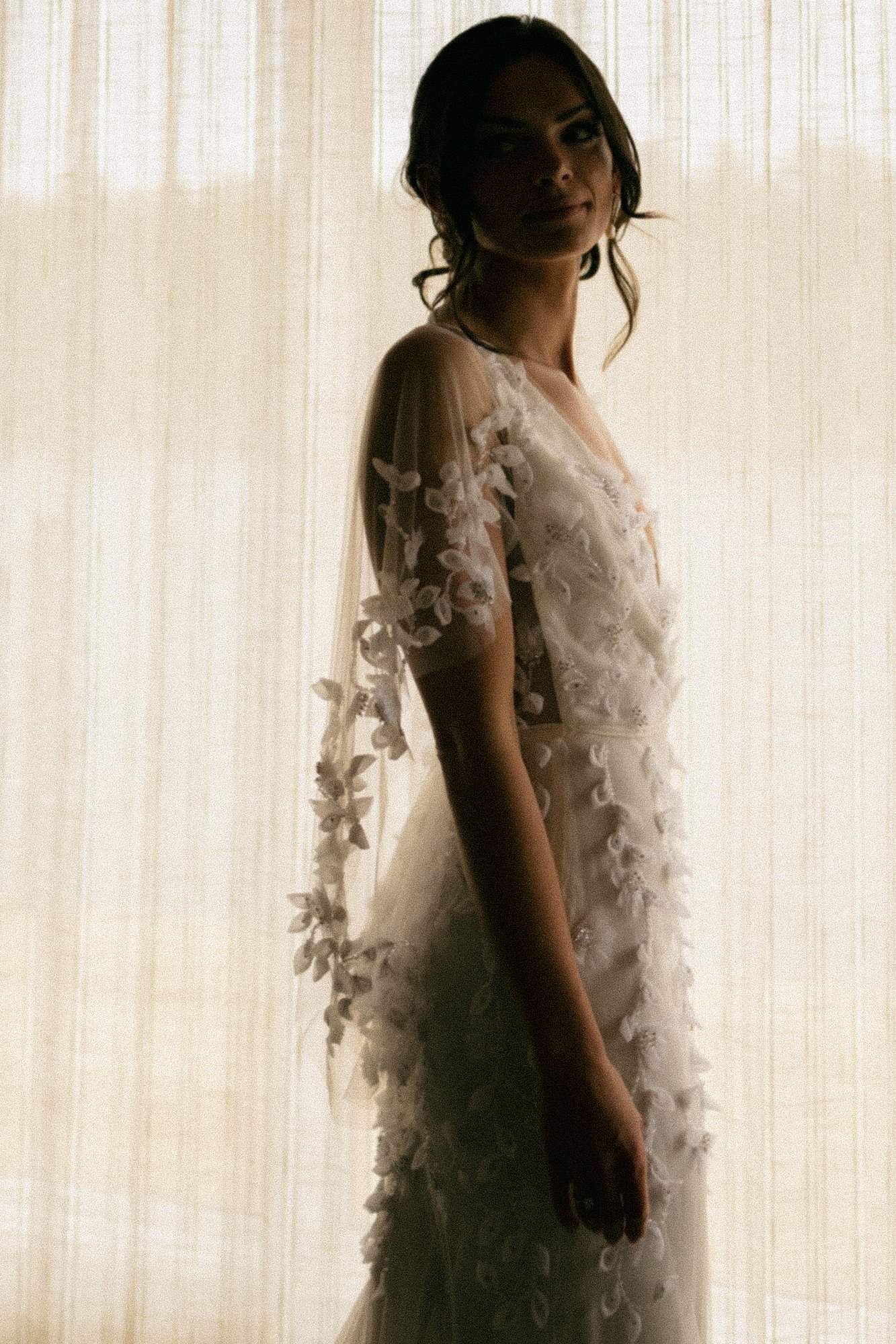 A woman in a white dress with floral embellishments, standing against a sheer curtain background, smiling softly and looking towards the camera.