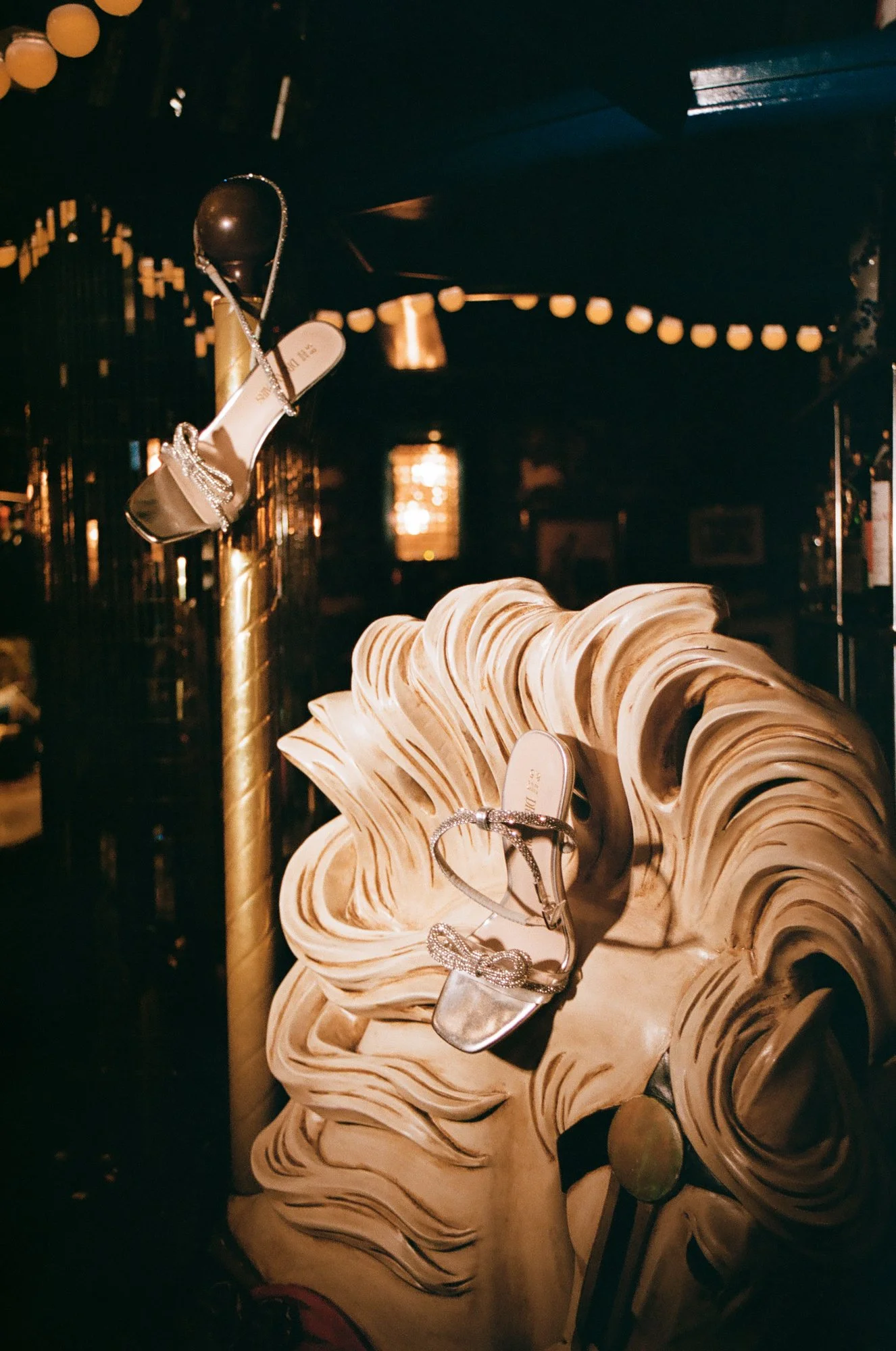 Display of women's wedding heels with rhinestones set on a carved carousel horse inside a dimly lit restaurant bar with string lights overhead.