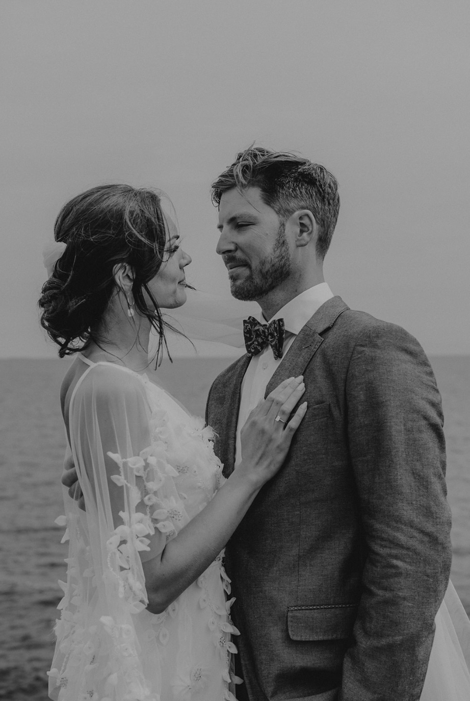 A black and white photo of a bride and groom facing each other outdoors, with the ocean in the background. The bride has dark hair styled up with loose waves and wears a dress with flower details. The groom has short hair, a beard, and wears a suit with a bow tie. They are close, looking into each other's eyes, with the bride's hand on the groom's chest.