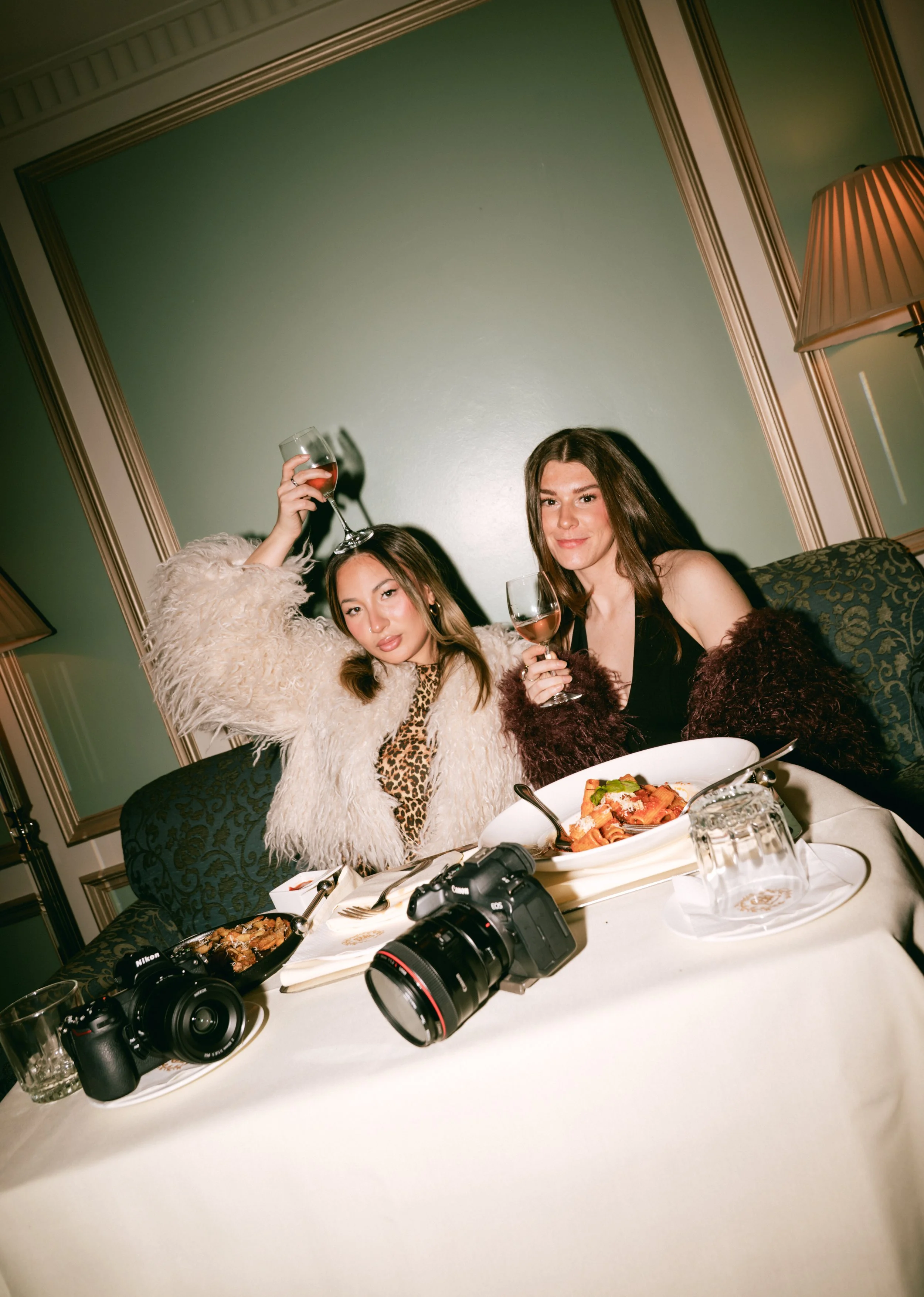 Two women celebrating at a dinner party, holding glasses of red wine, sitting at a table with food, cameras, and glasses, in a room with green walls and a lamp