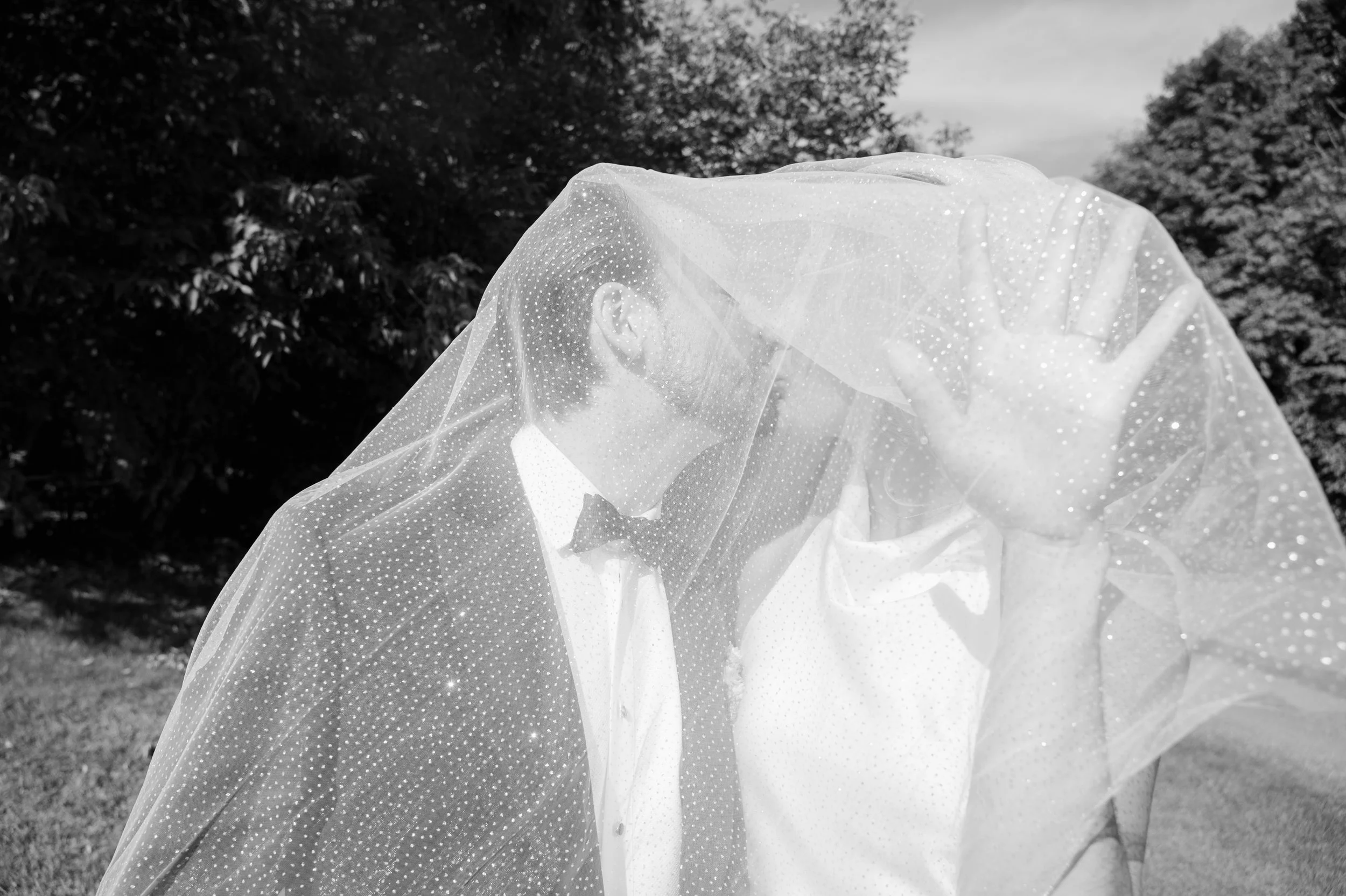 Black and white photo of a bride and groom under a veil outdoors with trees in the background.