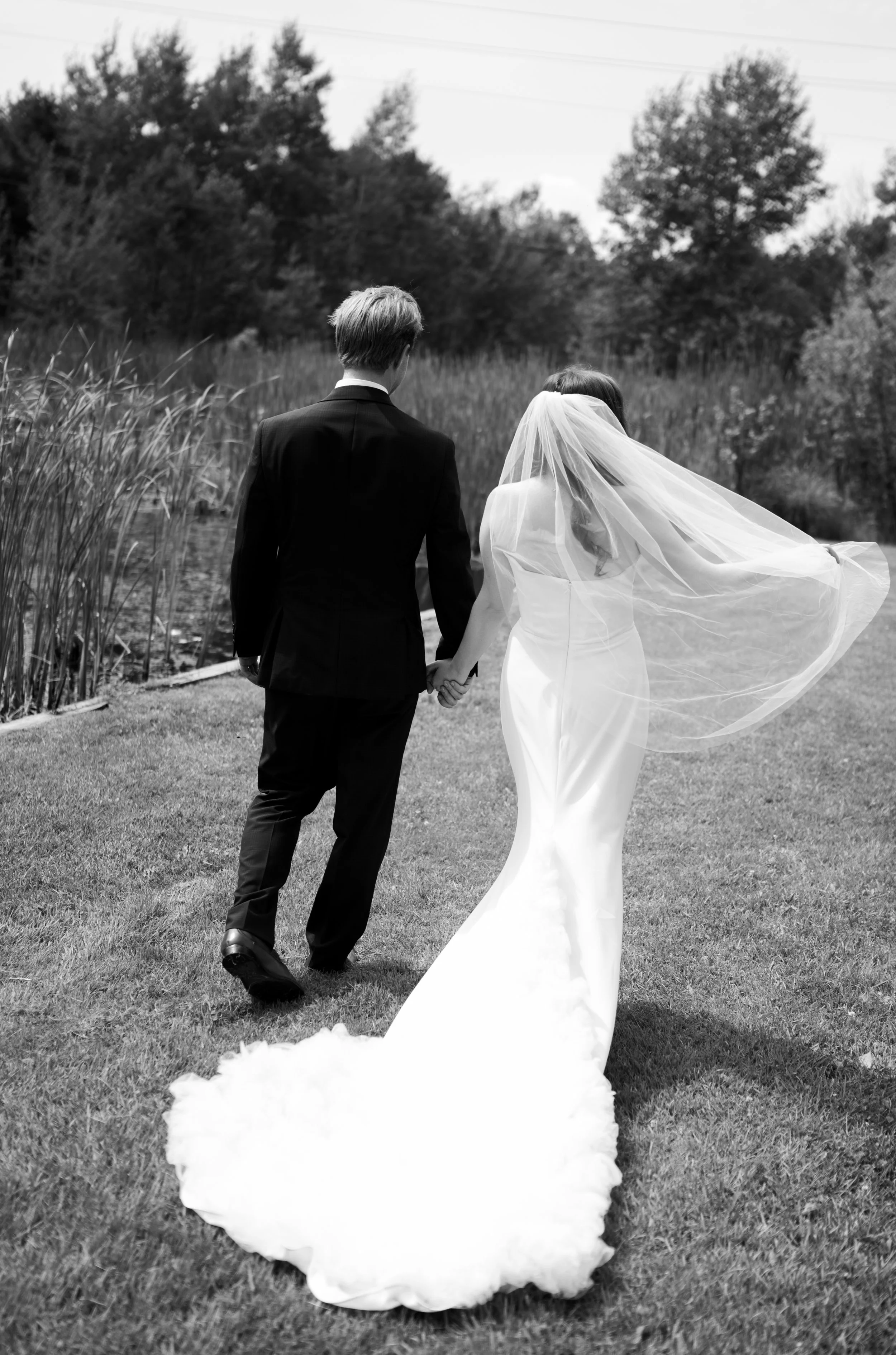 A black and white photo of a newlywed couple holding hands and walking away from the camera in a grassy outdoor setting. The bride is wearing a long satin wedding dress and veil, while the groom is dressed in a black suit. Trees and power lines are v