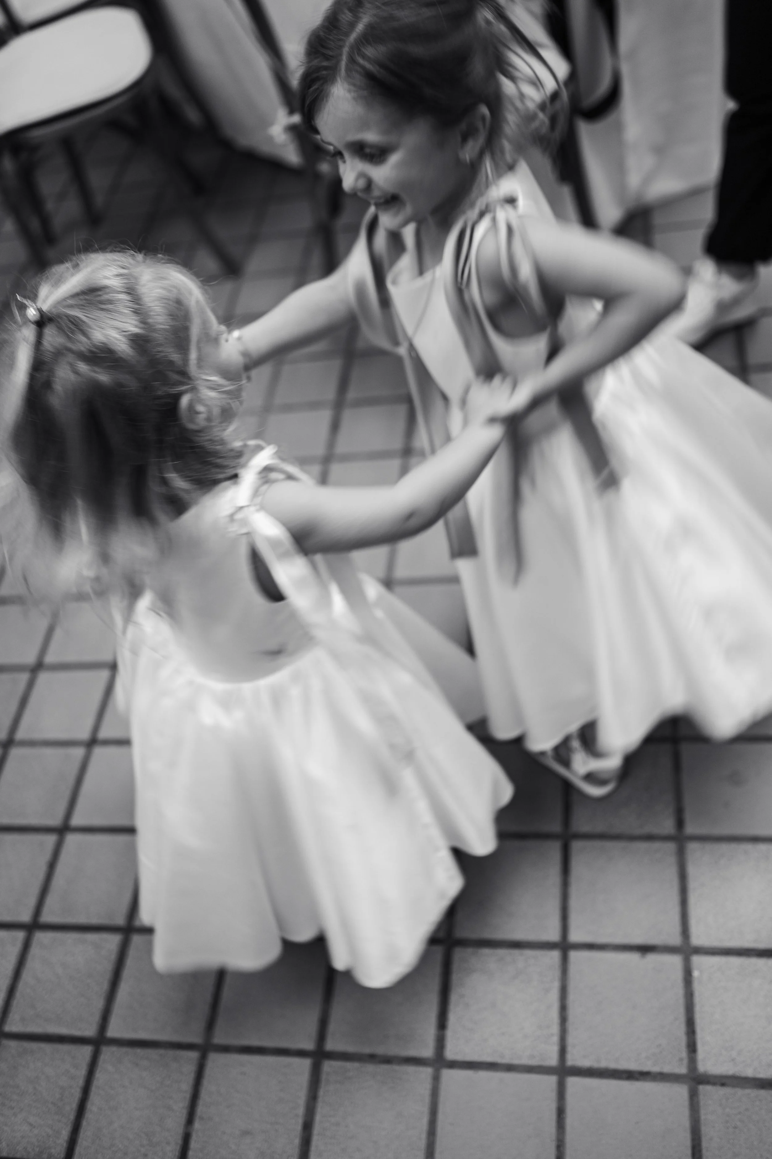 Two young girls in dresses, one with long hair in a ponytail and the other with shoulder-length hair, are holding hands and dancing together on a tiled floor. The girl with long hair is looking up at the other girl, smiling.