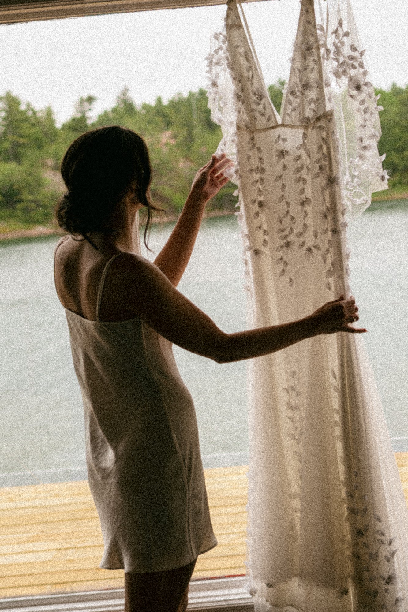 A woman standing by a window, adjusting curtains, with water and greenery visible outside.