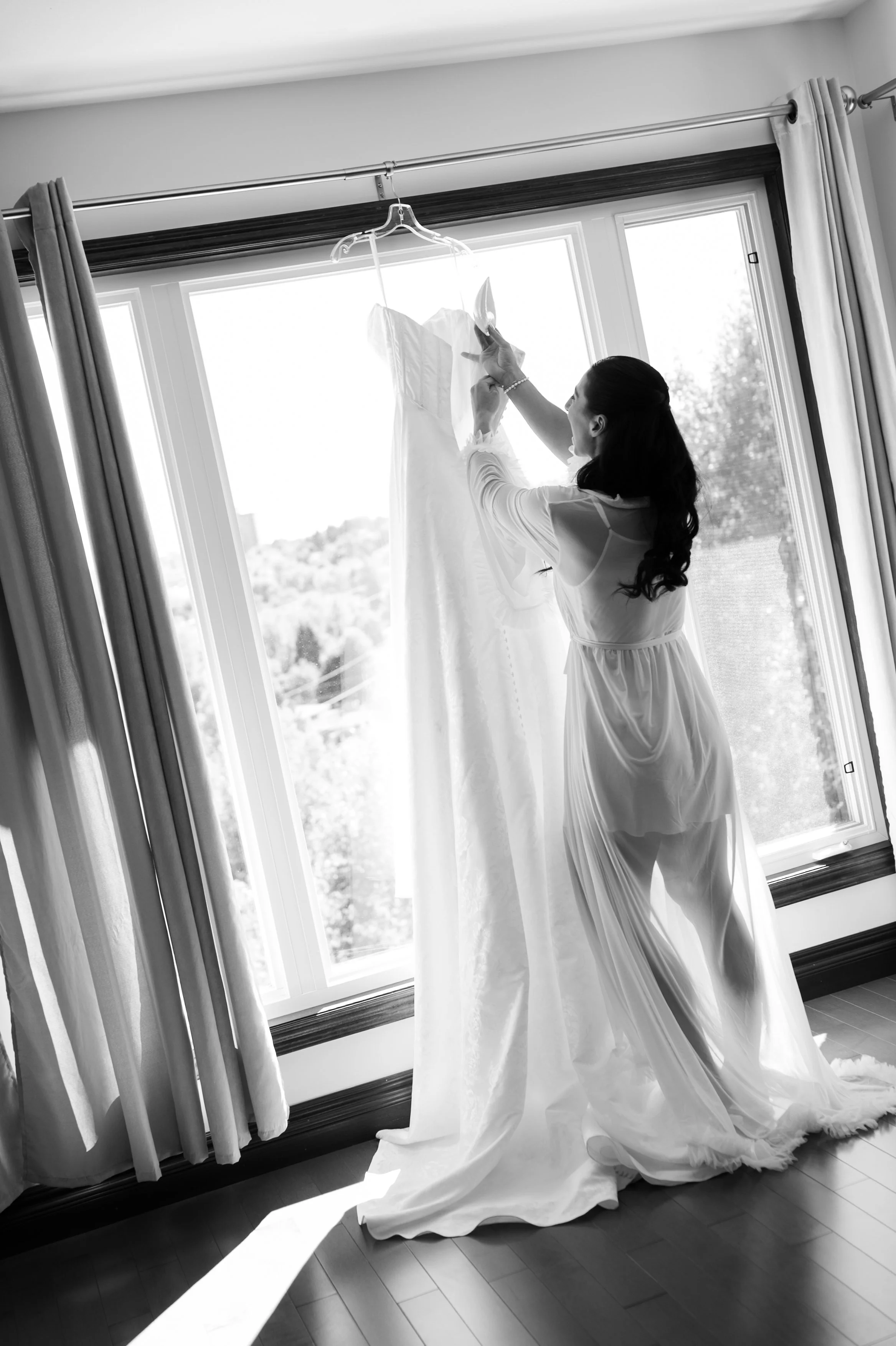 A woman with long dark hair wearing a white satin dress, viewed from the side, stands by a large window with sunlight streaming in. She is adjusting a wedding dress hanging on a clear hanger in front of her, with curtains on either side of the window