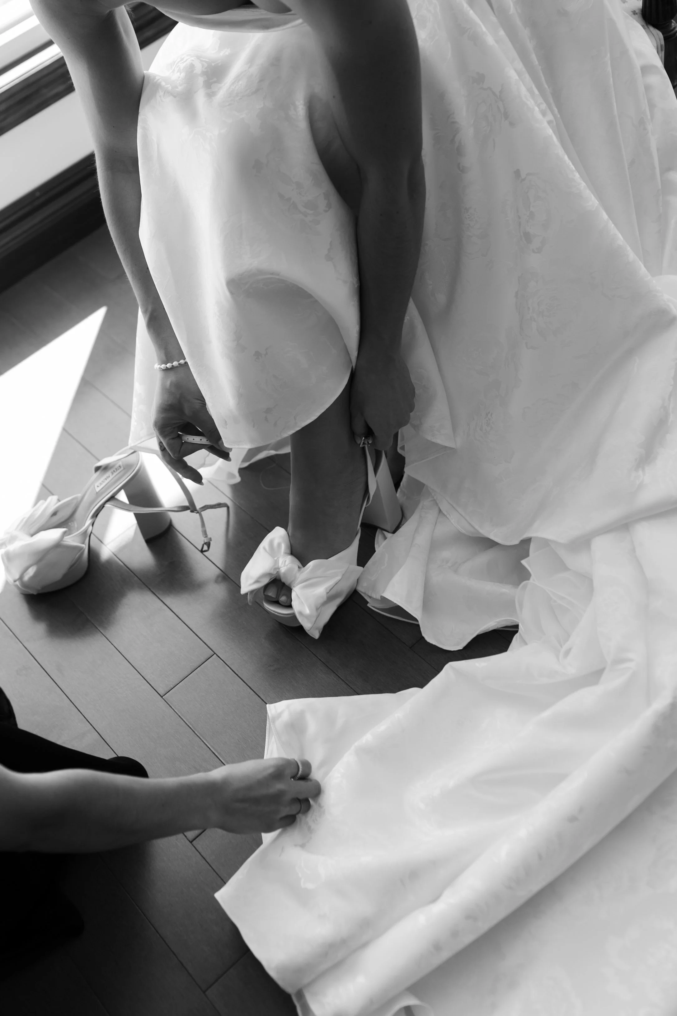 Black and white photo of a bride sitting on the floor, wearing a wedding dress and holding a piece of jewelry, with her legs crossed at her ankles, next to a window.