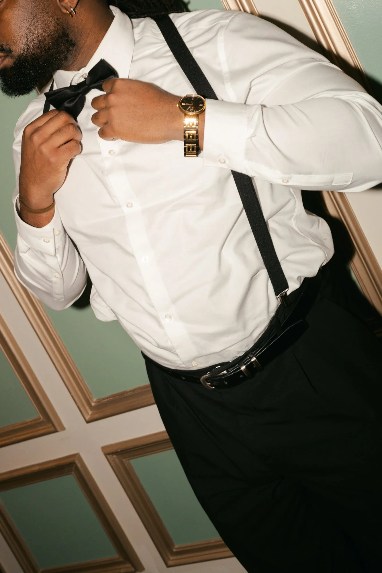 A man is seen adjusting a black bow tie while wearing a white dress shirt, black suspenders, a gold watch, and a black belt. 