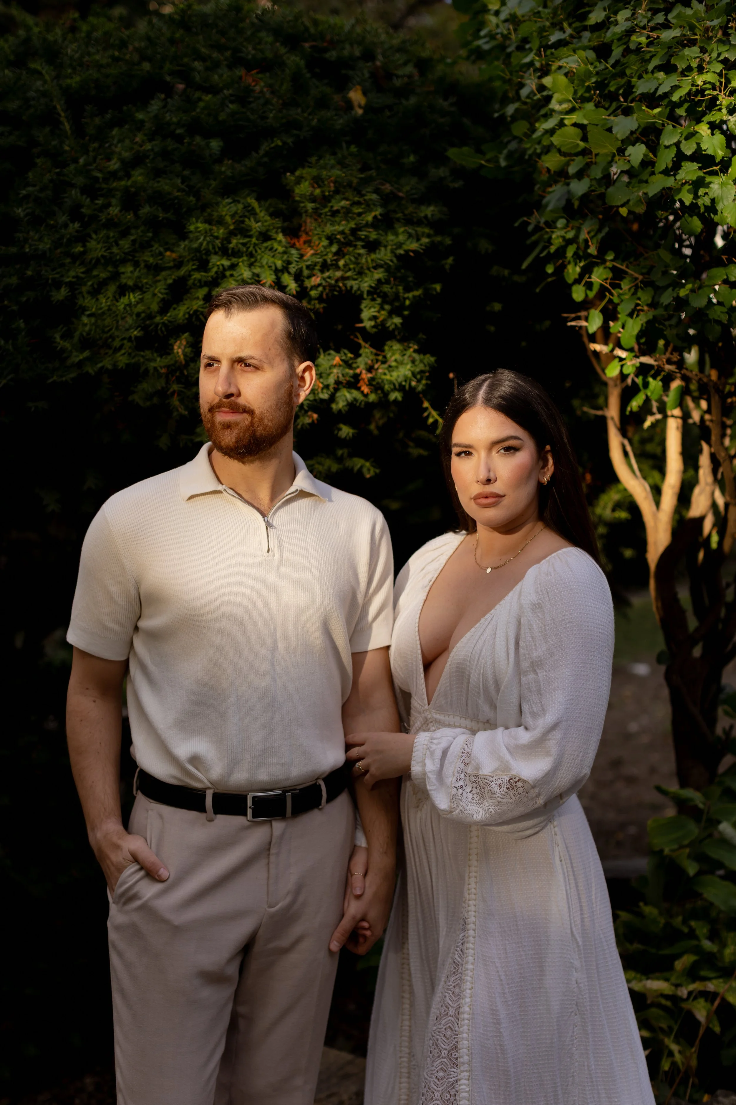 A man with a beard and a woman with dark hair standing outdoors in front of green foliage, holding hands.