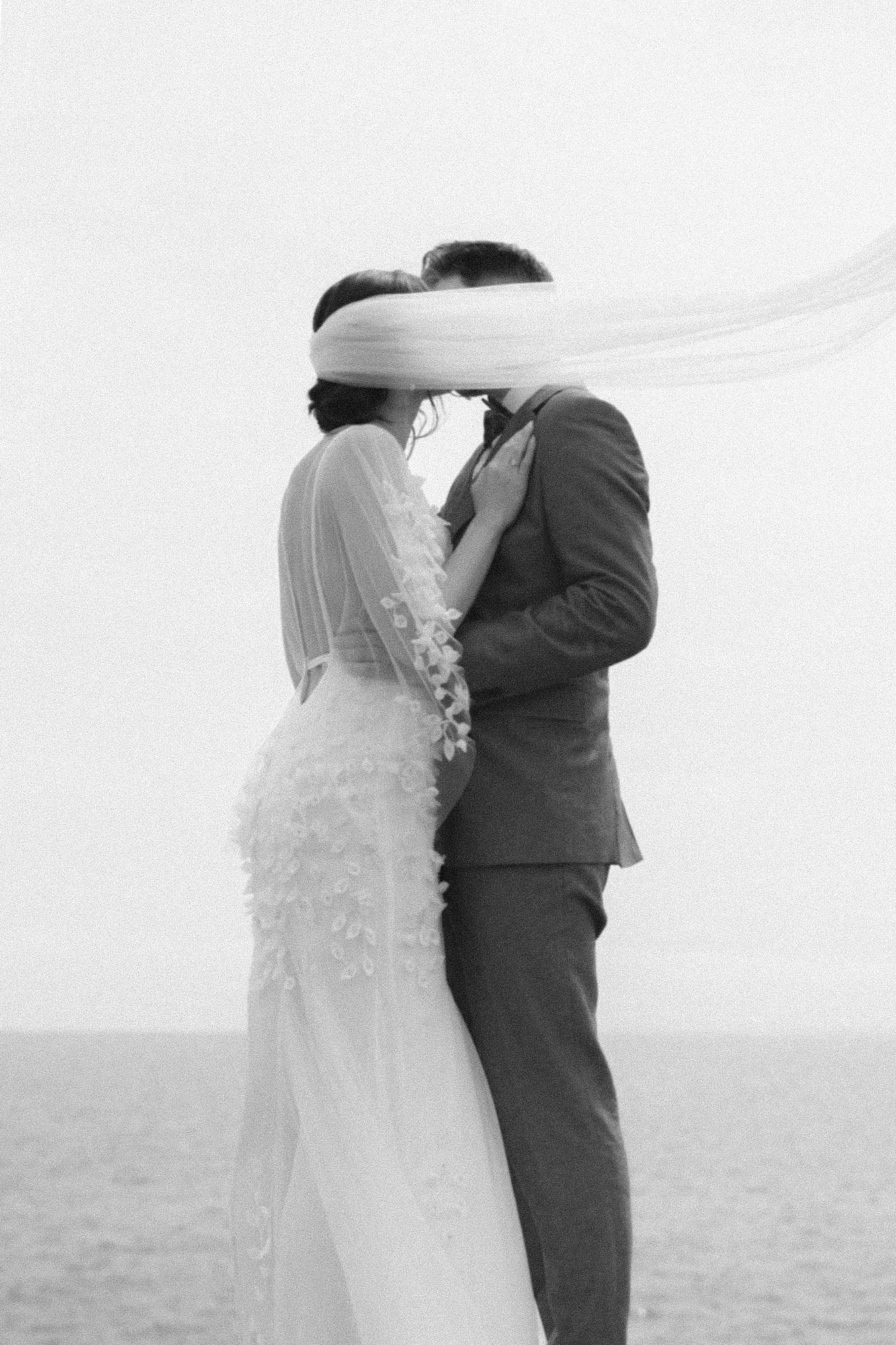 Black and white photo of a couple during their wedding ceremony, standing close and kissing on the beach. The bride is wearing a lace wedding dress with floral details, and the groom is in a suit. Their faces are covered with a flowing fabric in the air.