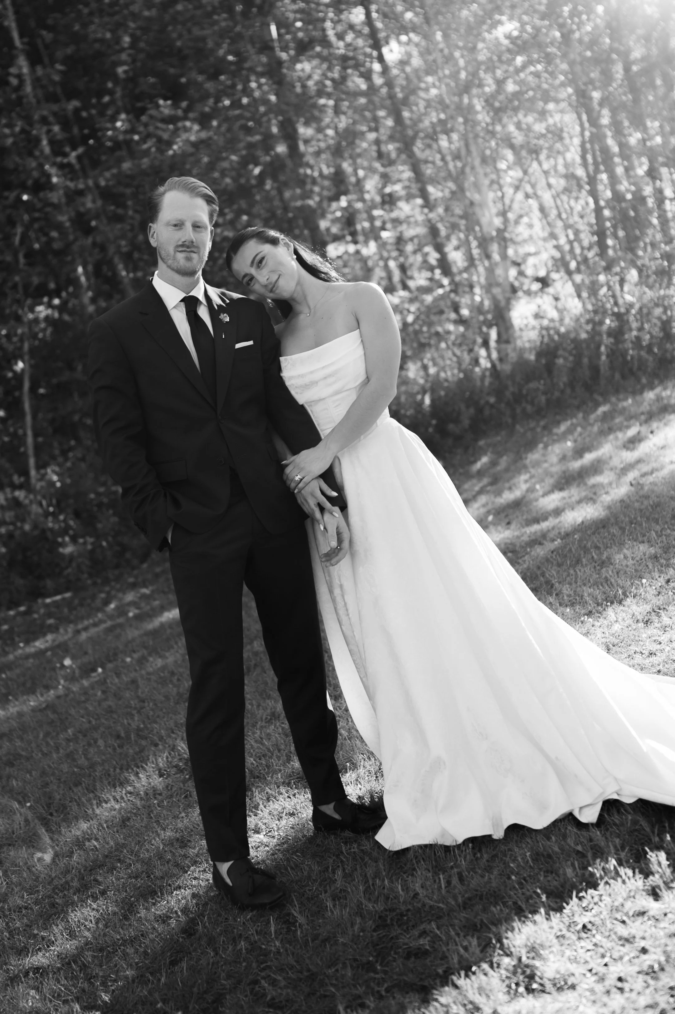 A black and white photo of a smiling bride and groom standing outdoors, with the bride resting her head on the groom's shoulder. They are dressed in wedding attire, and the background features trees and sunlight.