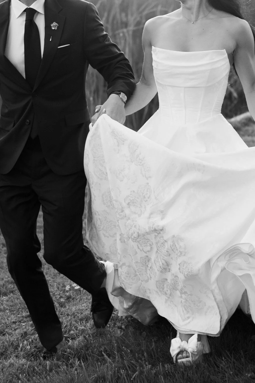 A bride in a white wedding dress and a groom in a black suit holding hands and walking outdoors.