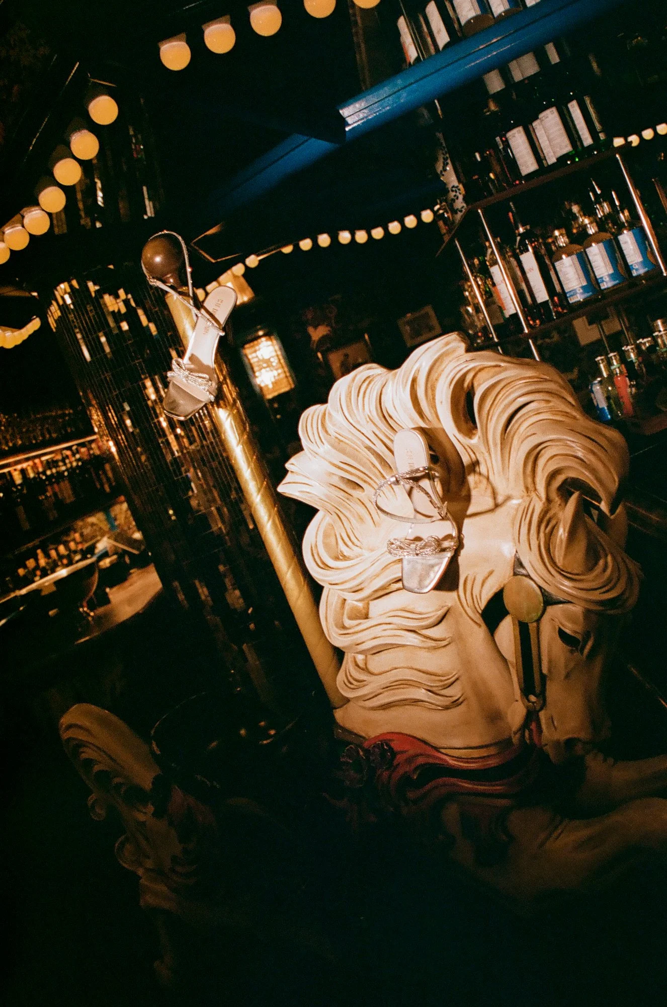 Display of women's wedding heels with rhinestones set on a carved carousel horse inside a dimly lit restaurant bar with string lights overhead.