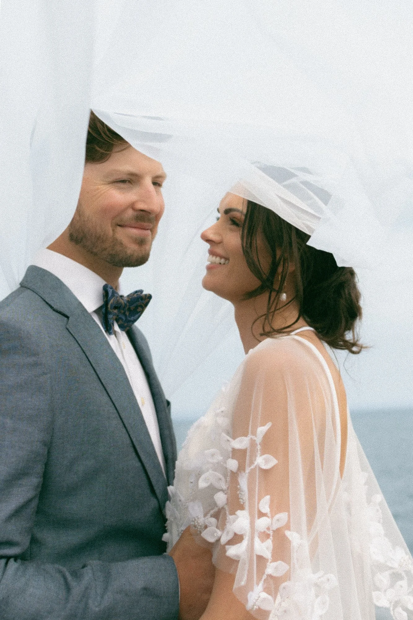 A bride and groom standing closely together under a sheer white veil, smiling at each other outdoors.