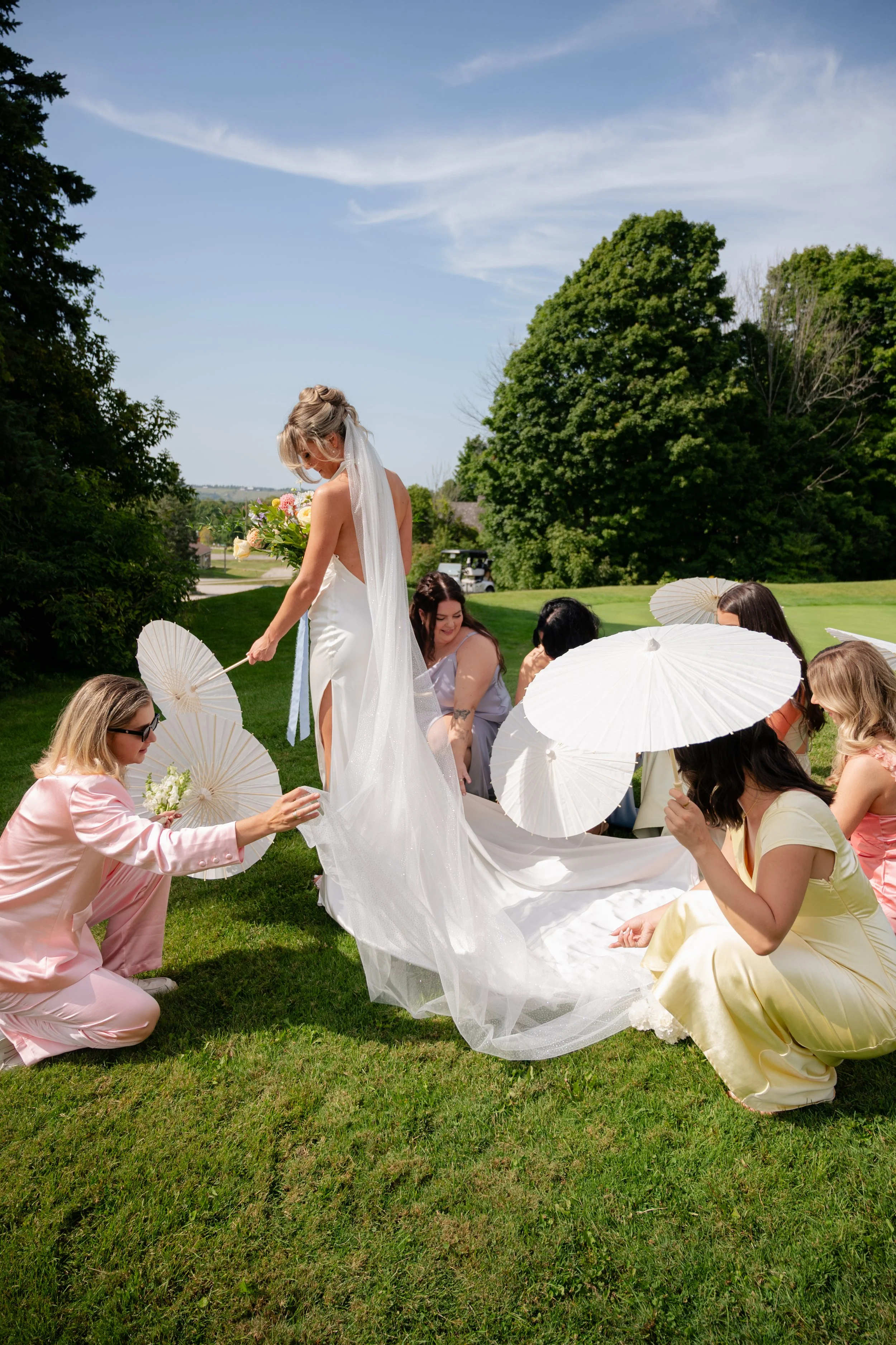 A group of women gather outdoors on a grassy field during a wedding celebration, with a bride in a white wedding dress and veil at the center, holding a bouquet, among guests holding white parasols, with trees and blue sky in the background.