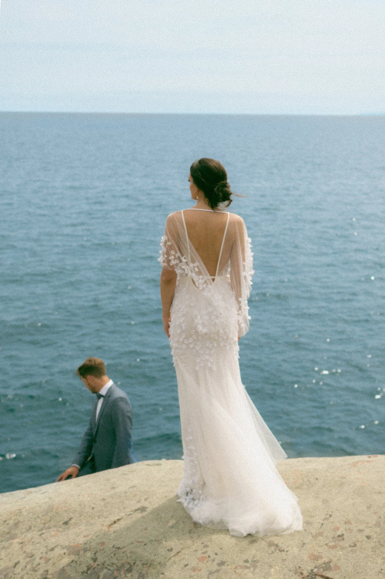 A woman in a white wedding dress standing by the ocean, with two men in suits below her, one looking down and the other walking away.