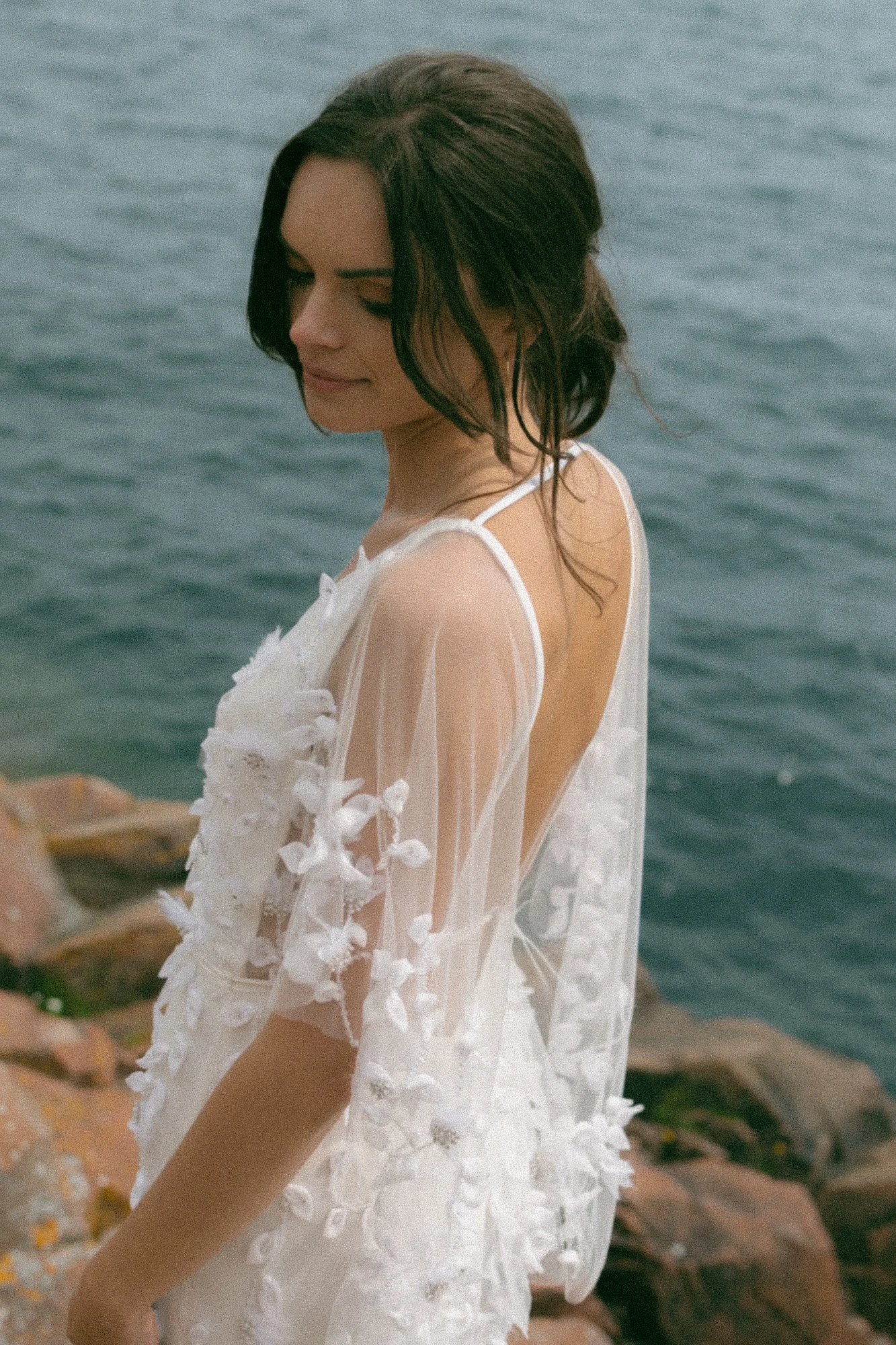 A young woman with dark wet hair and light skin posing outdoors near water, wearing a sheer white dress with floral appliqués.