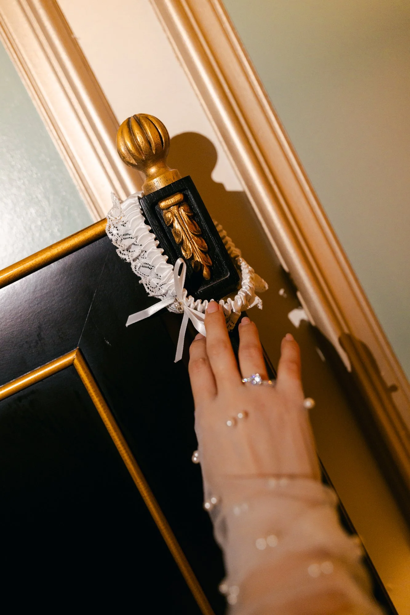 A person's hand with a pearl bracelet and ring touches a black wall with gold decorative trim, topped by a gold finial on a black and gold piece of furniture or decor, in a room with beige molding and a green wall.