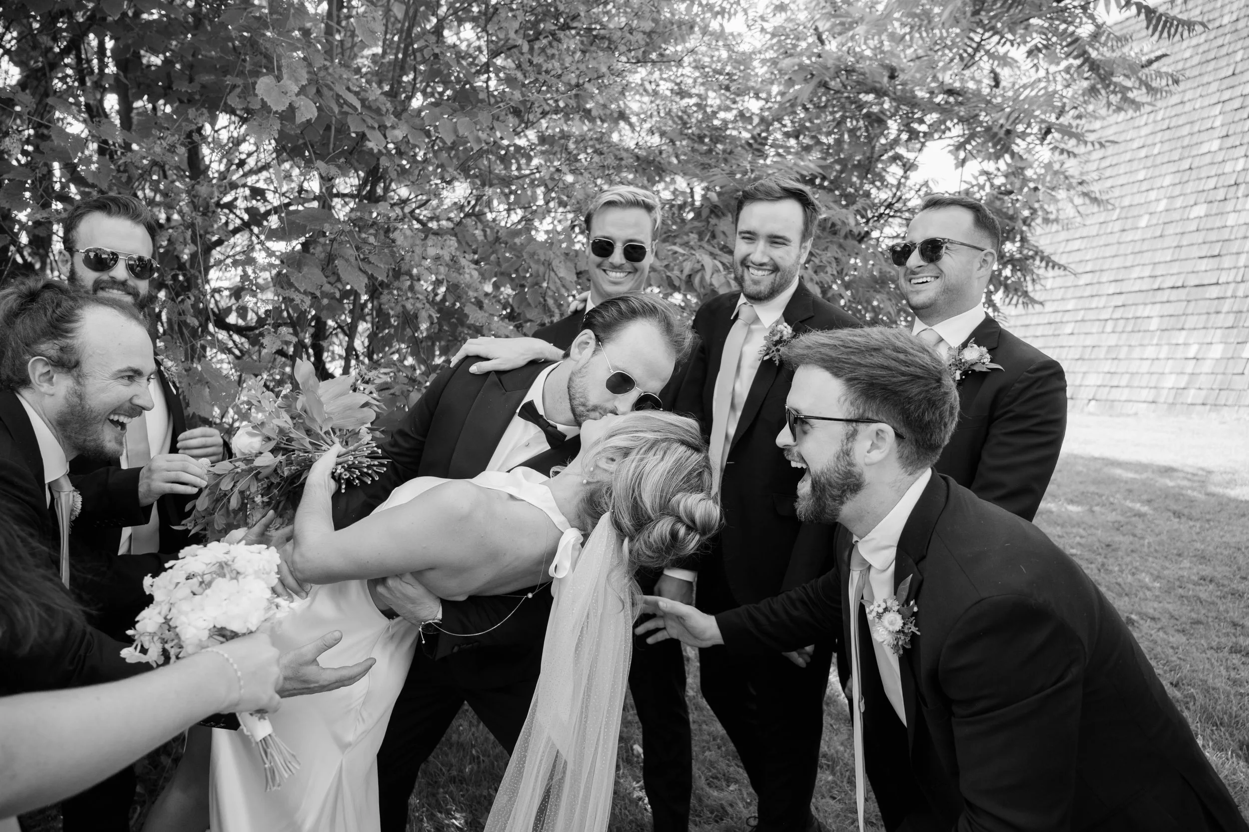 Black and white photo of a bride and groom leaning in to kiss, surrounded by friends and family in tuxedos and dresses, all smiling and laughing outdoors near a brick wall and trees.