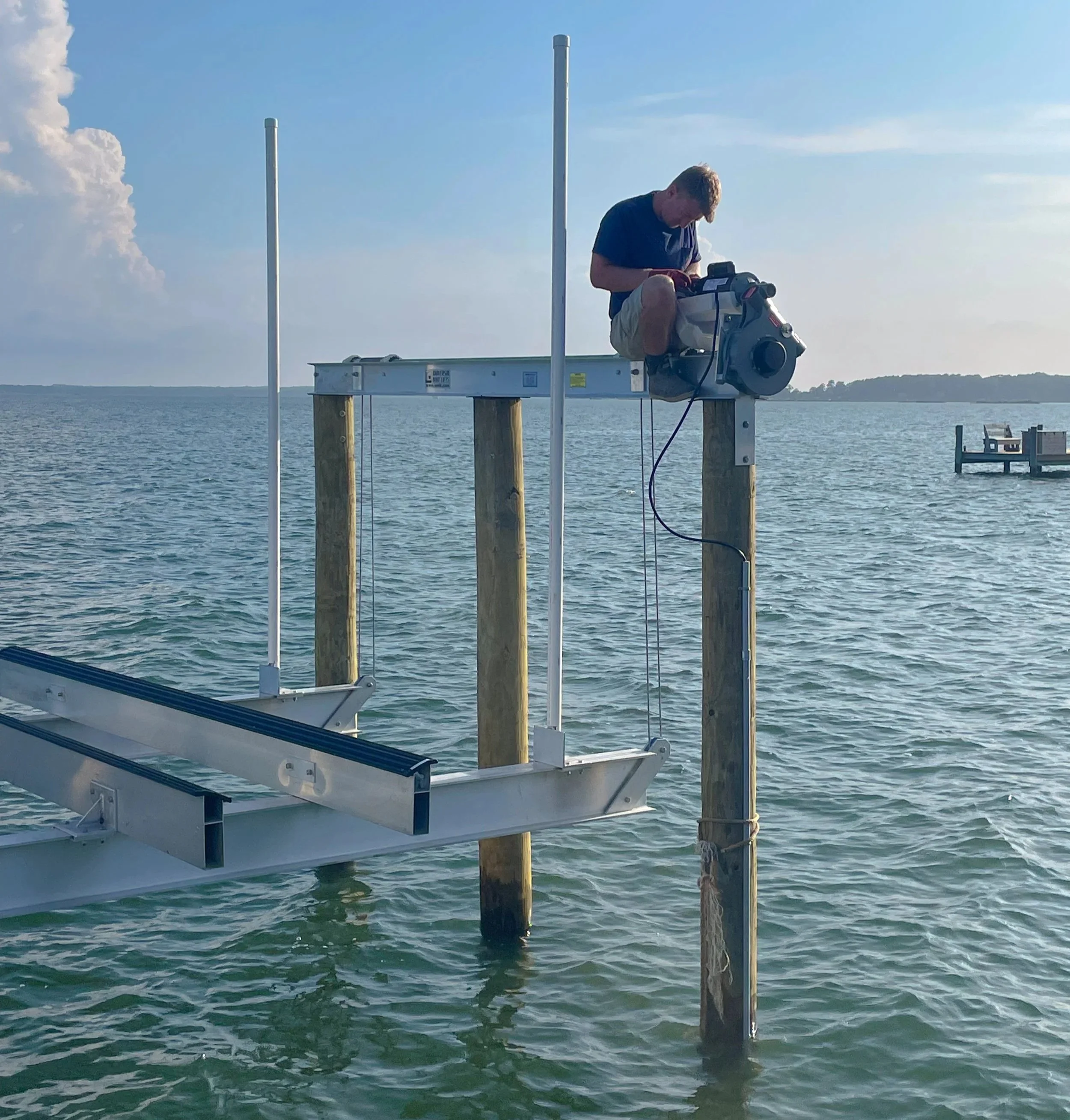 A Keeners Electric employee working on a boat dock by the water, adjusting or installing equipment on wooden posts, with the ocean and a distant shoreline in the background.