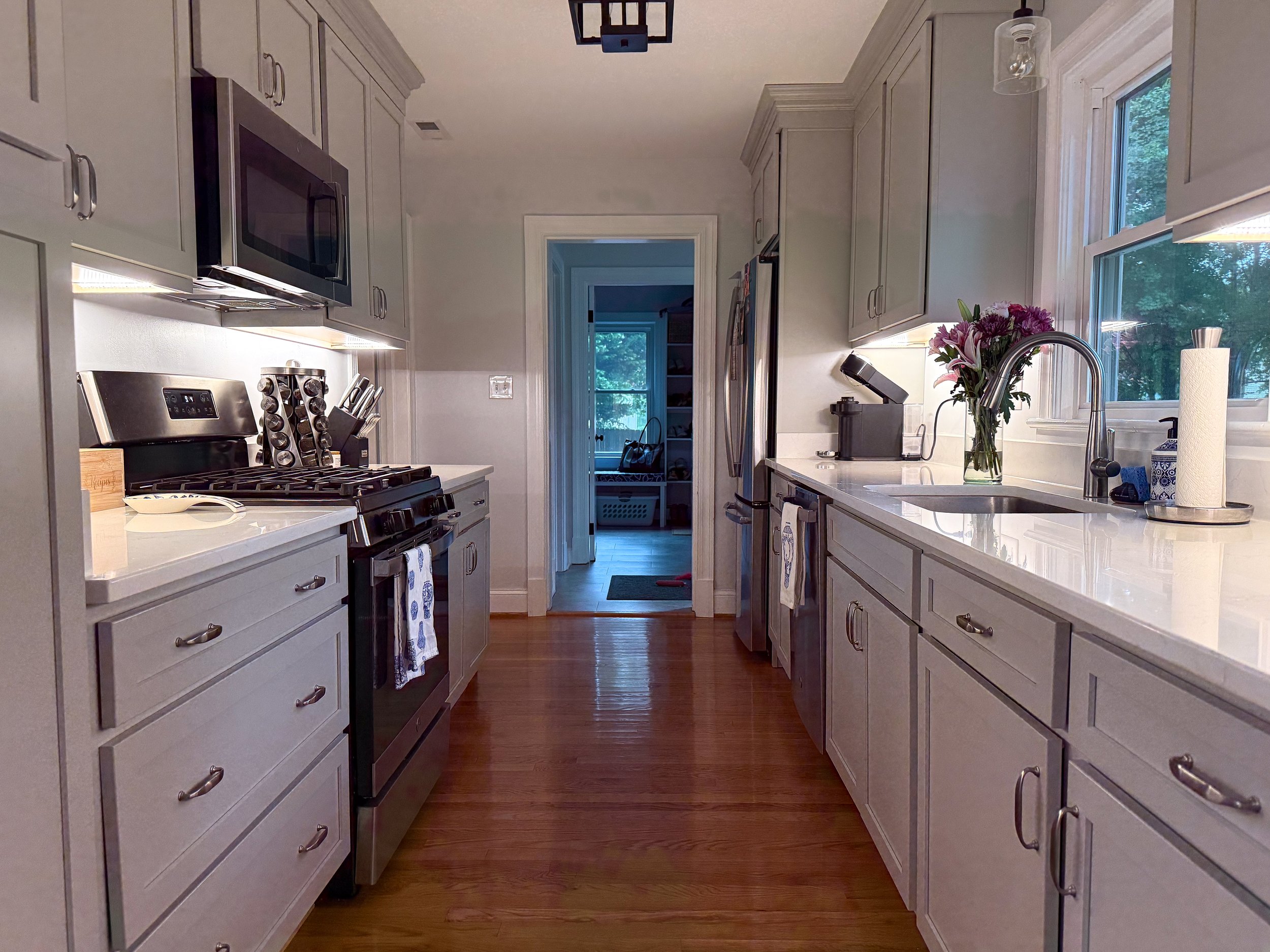 A modern kitchen with white cabinets, a white countertop, a stainless steel stove, microwave, and refrigerator. There are flowers in a vase on the counter, a coffee maker, and a window letting in natural light. An open doorway leads to another room.