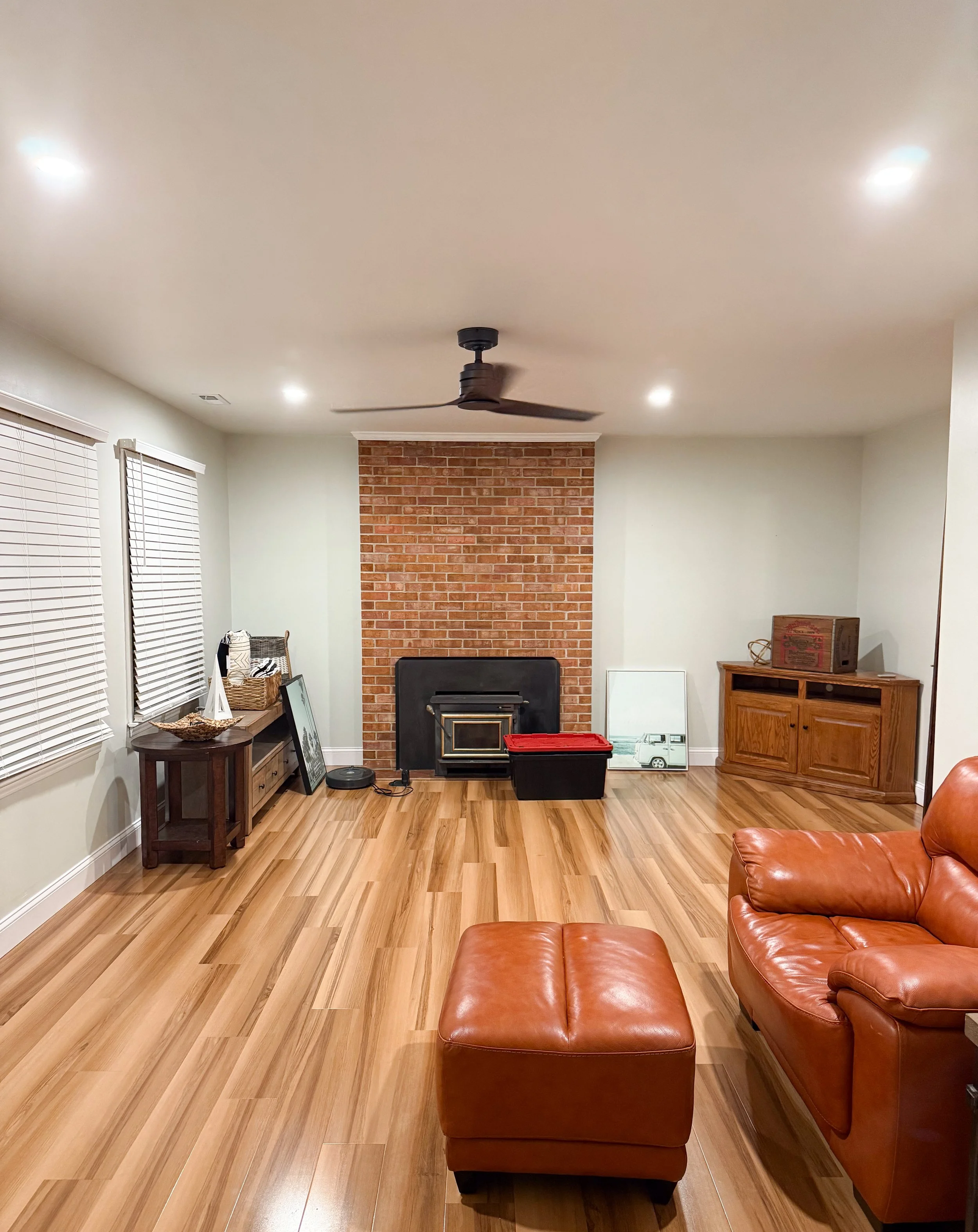 Living room with wooden floor, leather sofa, ottoman, brick fireplace, ceiling fan, and windows with blinds.