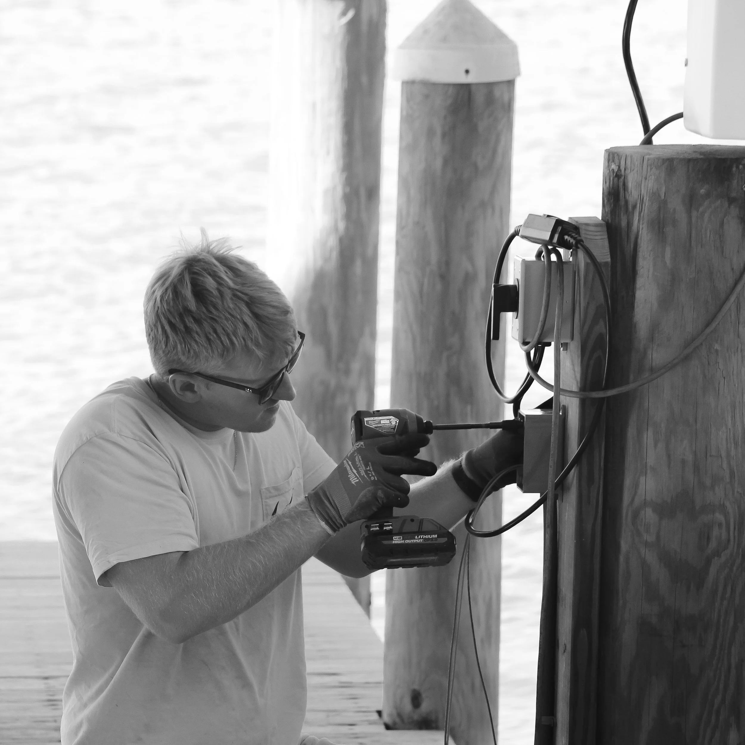 A Keeners Electric employee wearing glasses and gloves is using a power drill on a wooden post installing electrical equipment.