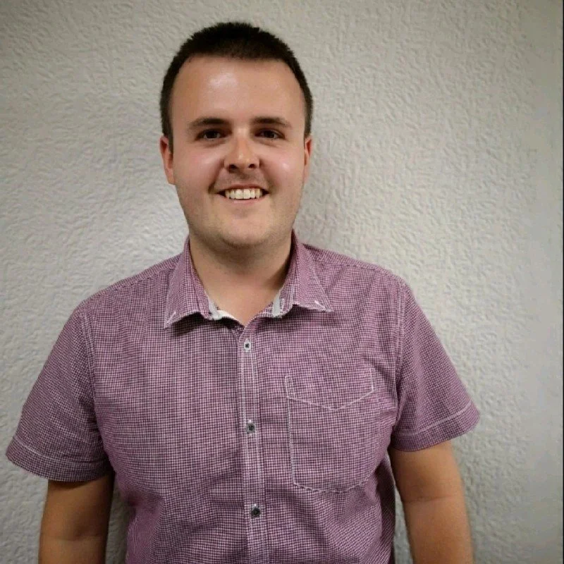 A young man with short dark hair and a big smile, wearing a short-sleeved, button-up, red checkered shirt, standing against a textured beige wall.