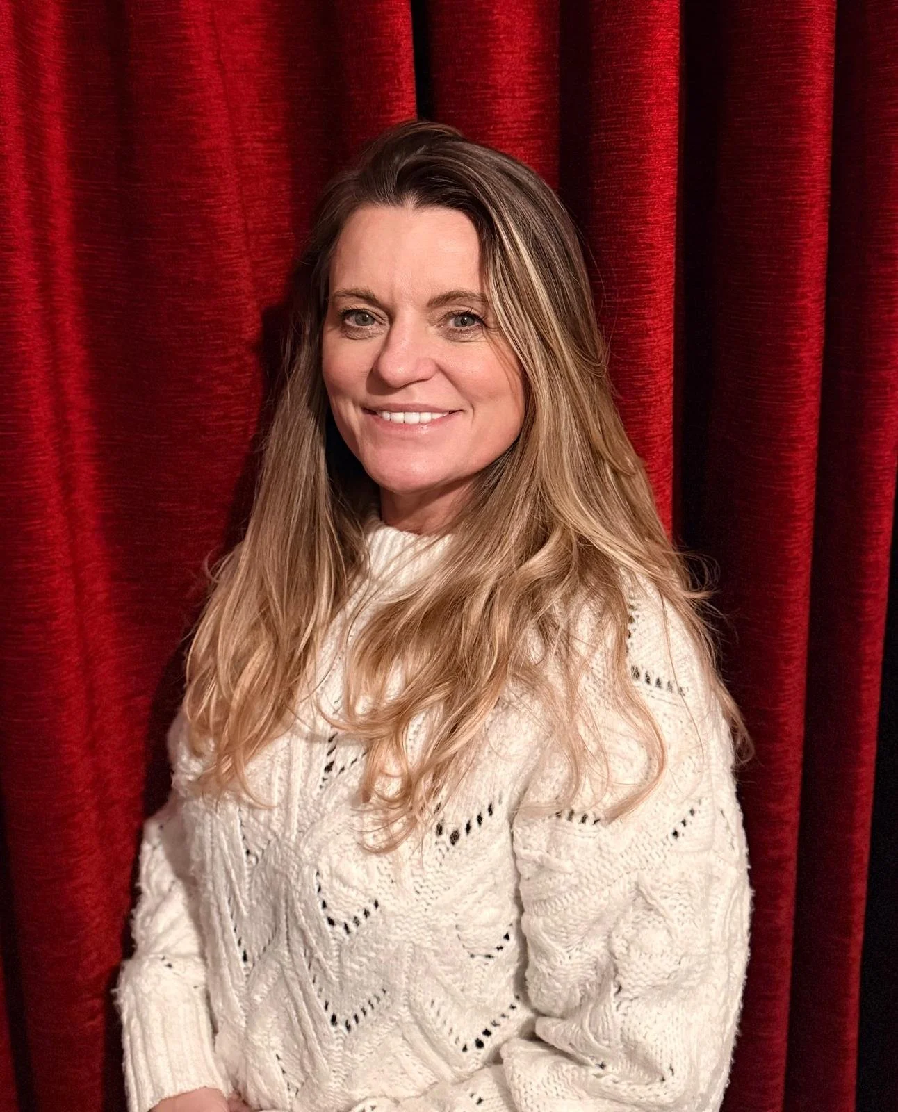 A woman with long, wavy blonde hair smiling in front of red velvet curtains, wearing a white knitted sweater.