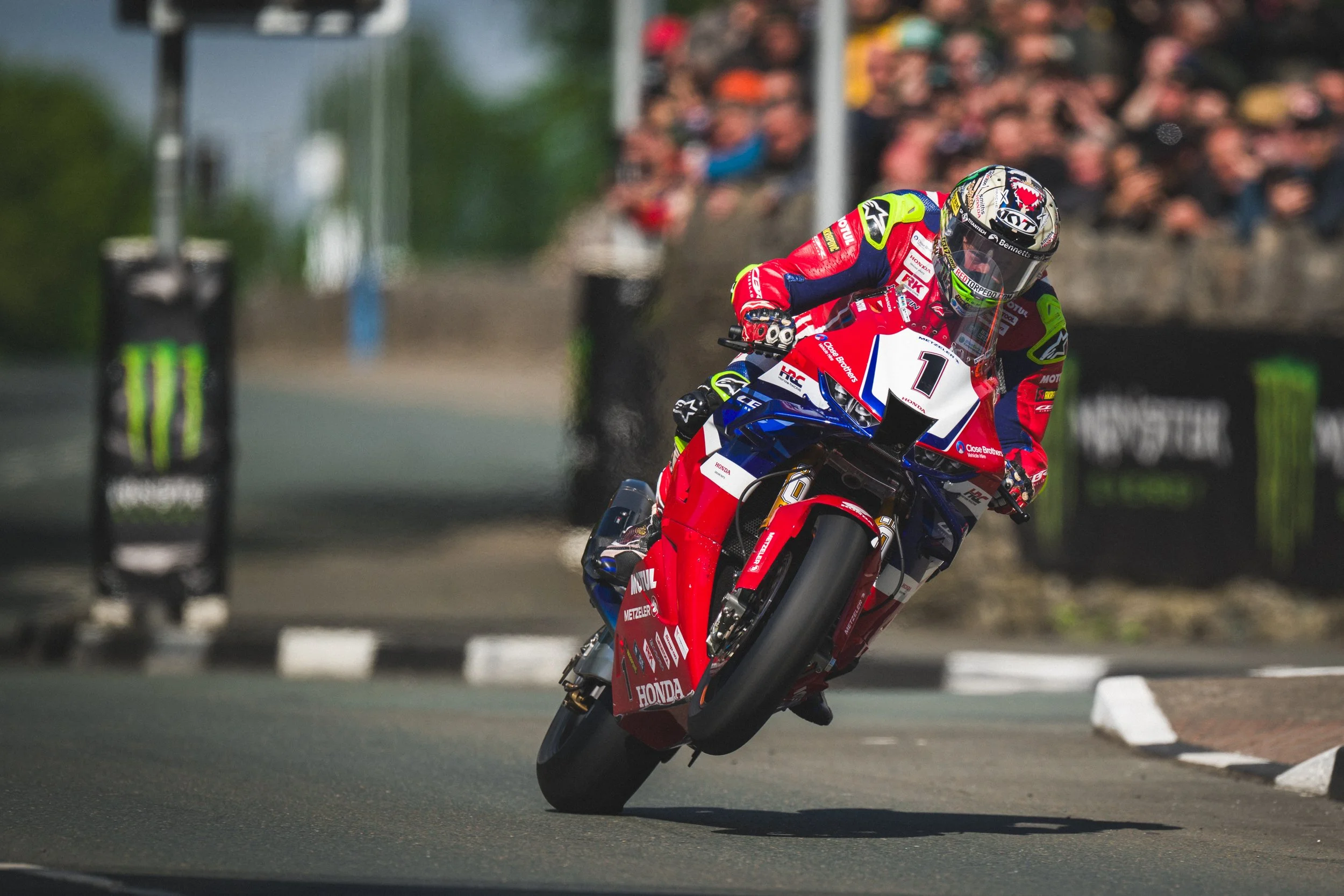 A professional motorcycle racer in a red, white, and blue racing suit leaning into a turn on a racetrack, riding a red and blue Honda motorcycle with the number 1 displayed on the front, while a crowd of spectators watches in the background.