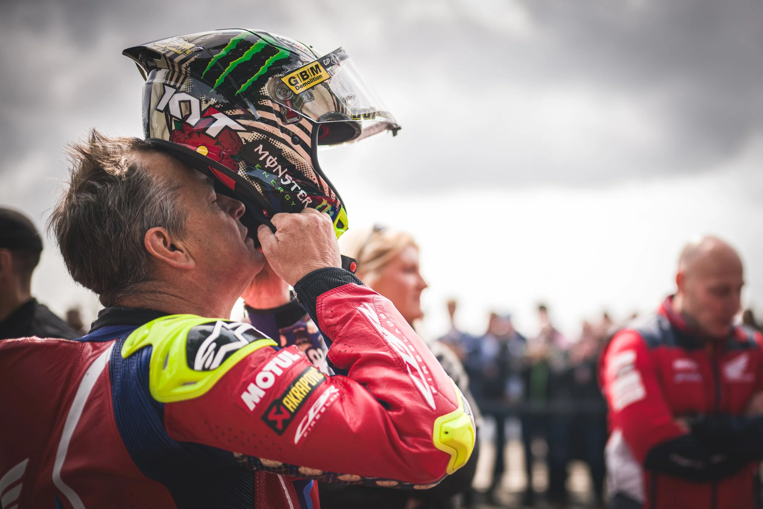 A man in a red racing suit holding a motorcycle helmet with a sky and a crowd in the background.