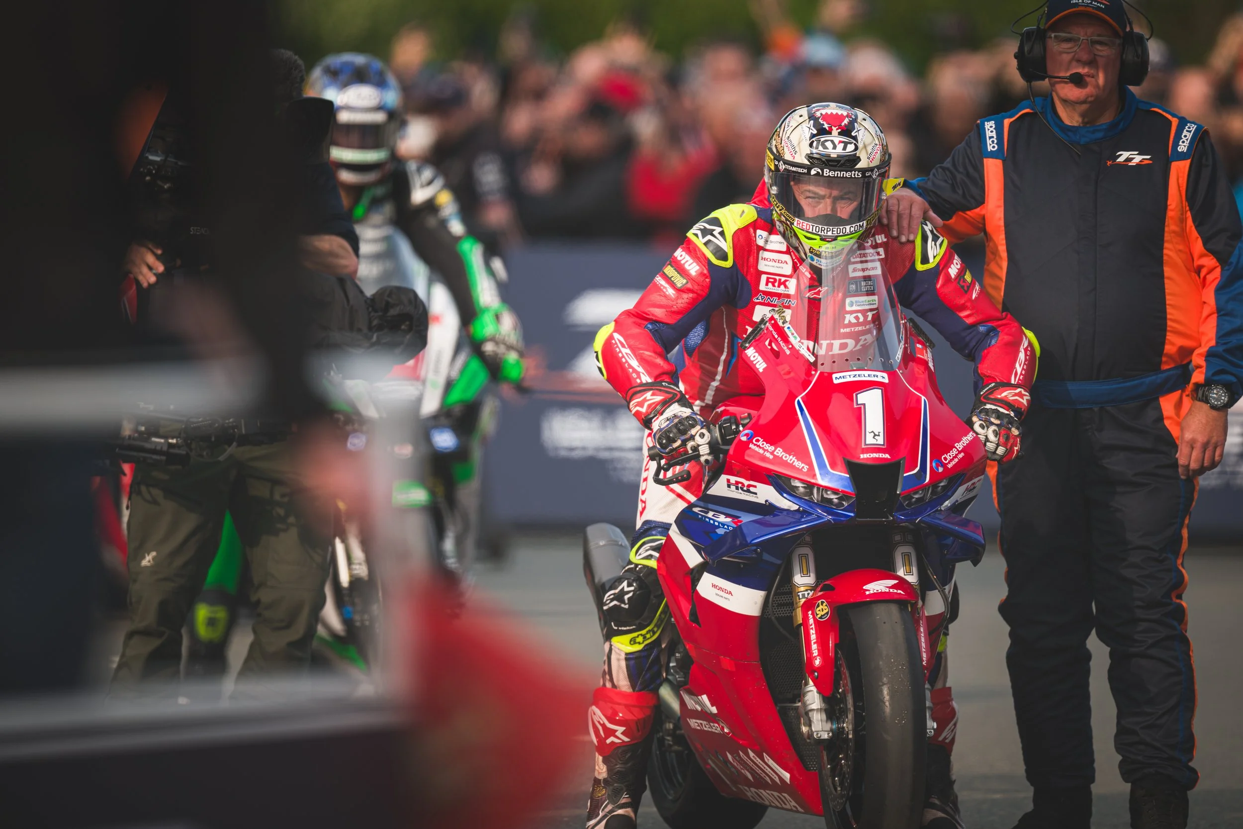 A motorcyclist in red and blue racing gear and helmet sitting on a Honda race bike, with team staff standing beside in a race paddock, and other racers and spectators in the blurred background.